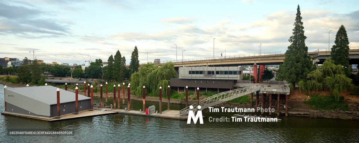 Portland Fire & Rescue Station 21 sits serenely on the Willamette River beneath soft afternoon clouds, its modern floating dock structure anchored by rust-red pilings that pierce the calm waters. The sleek gray boathouse contrasts against the verdant landscape of Central Eastside, where towering evergreens frame the Madison Bridge's concrete span. Willow trees cascade toward the riverbank while urban infrastructure creates geometric layers in the background, capturing the harmonious intersection of emergency services and Portland's natural waterways.