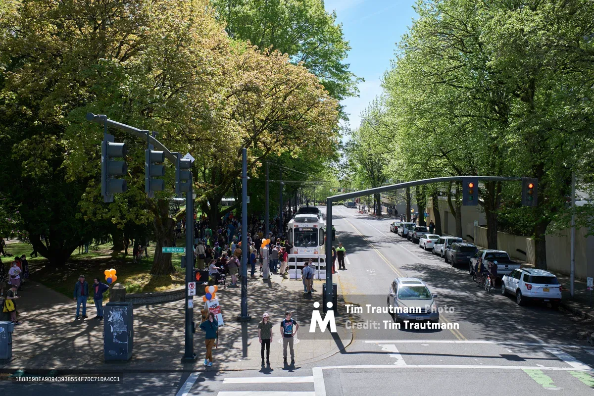 Crowds gather around a vintage white TriMet MAX Type 1 light rail car during its farewell ceremony at Holladay Park in Portland's Lloyd District. Dappled sunlight filters through mature spring foliage, casting dancing shadows across the assembled transit enthusiasts and officials in safety vests. The iconic train sits beneath traffic signals on the tree-lined street, marking the end of an era for Portland's original light rail fleet. Orange balloons and ceremonial touches add warmth to this bittersweet urban transit milestone.