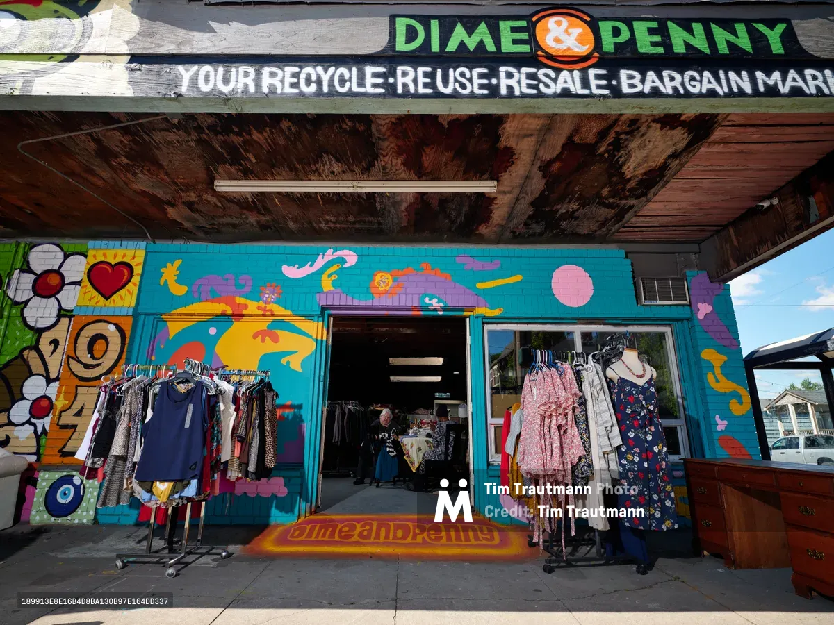 A vibrant thrift store facade on Northeast 15th Avenue in Portland's Alberta district showcases exuberant street art murals in turquoise, orange, and purple hues. Clothing racks spill onto the sidewalk beneath weathered wooden eaves, while colorful garments hang in the storefront windows. The hand-painted signage declares this a "recycle-reuse-resale-bargain mart," embodying the neighborhood's creative spirit and sustainable commerce ethos.