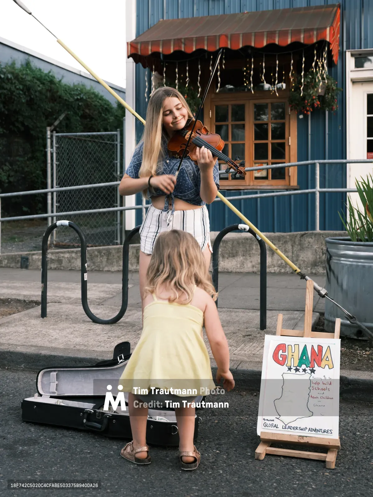 A blonde teenage violinist performs with passionate concentration at Portland's Last Thursday street fair on Alberta Street, her bow dancing across the strings as a young girl in a sunny yellow dress listens intently from beside an open violin case. The scene unfolds against the backdrop of Waffle Window's distinctive blue siding and red striped awning, with a handmade sign advertising a Ghana fundraiser completing this intimate moment of community connection through music. Golden hour light bathes the concrete plaza, transforming a simple street performance into a tender portrait of artistic sharing.
