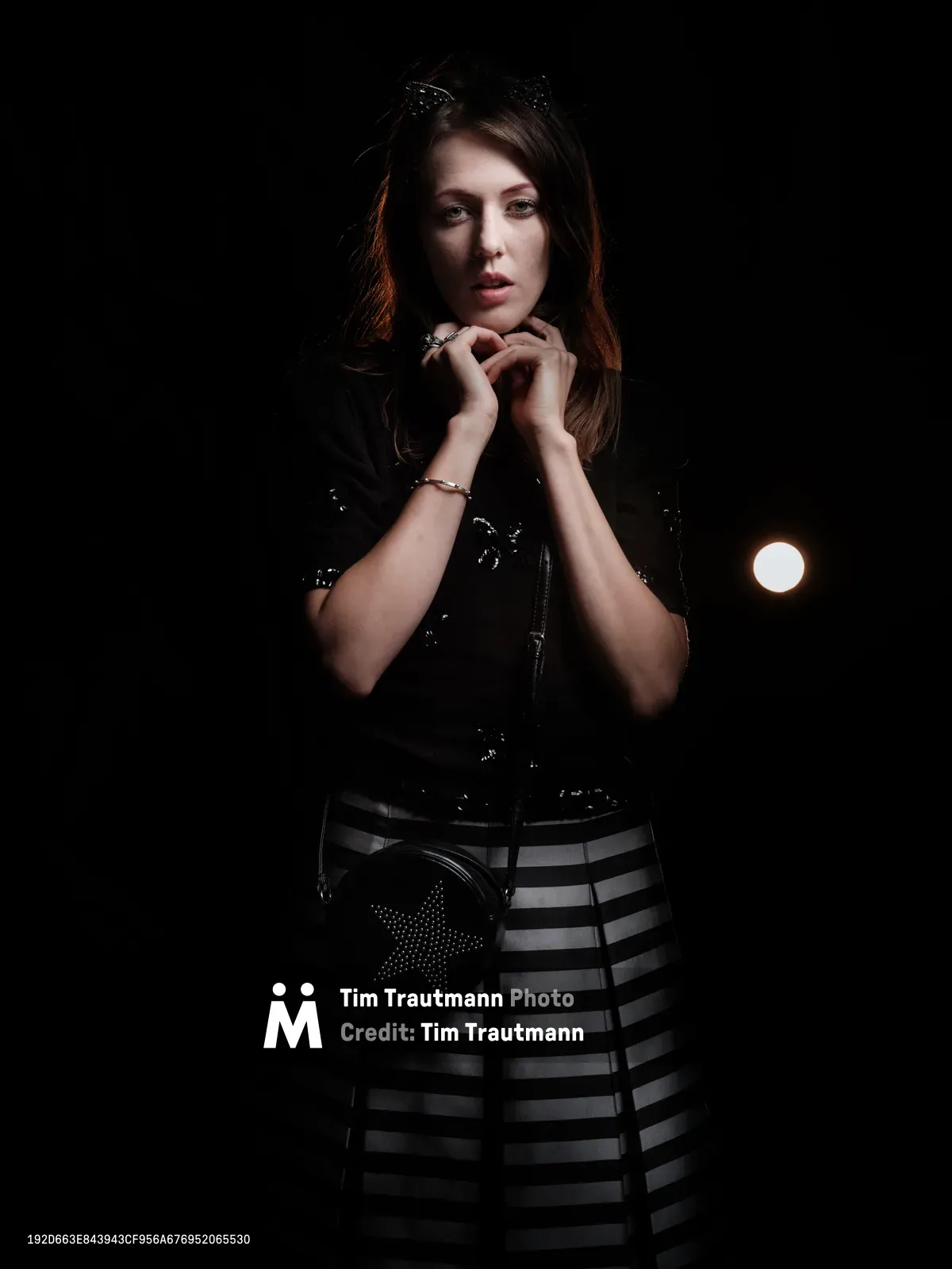 Rhianna Atwood emerges from deep shadows in this dramatic portrait captured in Portland's historic Kerns neighborhood. The chiaroscuro lighting sculpts her contemplative expression as she poses with hands clasped near her chin, wearing a black leather corset paired with a geometric striped skirt. A single warm light source pierces the darkness behind her, creating an intimate atmosphere of urban sophistication against the inky void.