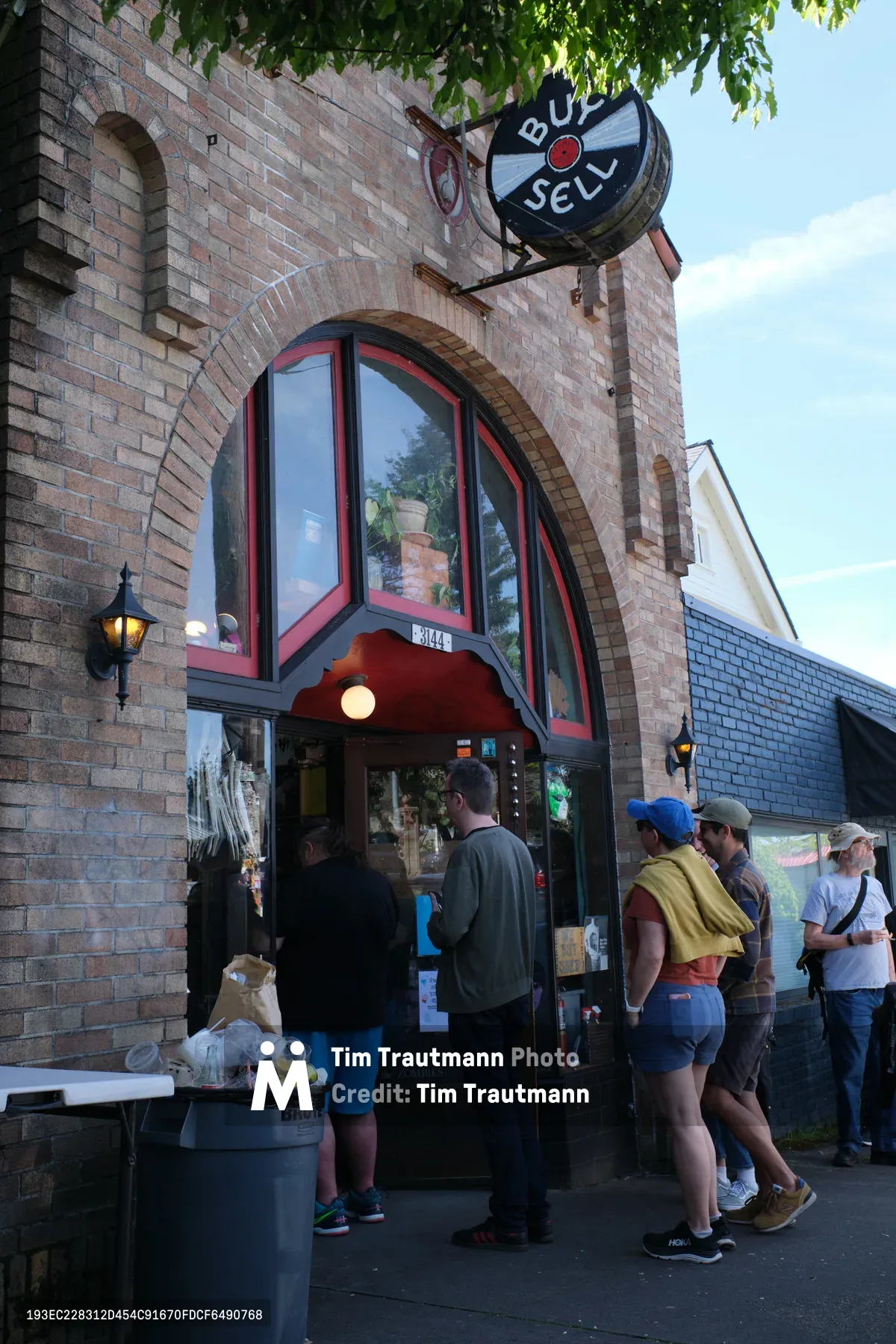 A diverse group of music enthusiasts gathers outside a distinctive brick record store in Portland's Kerns neighborhood, forming an orderly queue beneath the dappled shade of overhanging trees. The vintage architecture features striking arched windows with red trim and a prominent circular sign reading 'BUY SELL,' while soft afternoon light illuminates the weathered brick facade. Customers of various ages wait patiently, some carrying personal items, embodying the enduring culture of vinyl collecting and independent music retail that defines Portland's East Burnside corridor.
