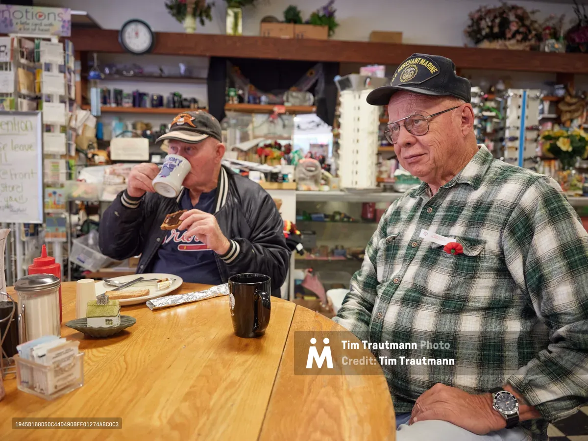 Two longtime Saint Johns residents, Jewel and Ron, share their regular morning ritual at Pattie's Home Plate Café on North Lombard Street. Jewel raises a ceramic mug while seated in a letterman jacket, as Ron in his plaid flannel and military cap sits beside him at their usual wooden table. The intimate neighborhood diner buzzes with the familiar comfort of decades-old friendships, surrounded by shelves lined with local memorabilia and artificial flowers that speak to the café's homespun character.