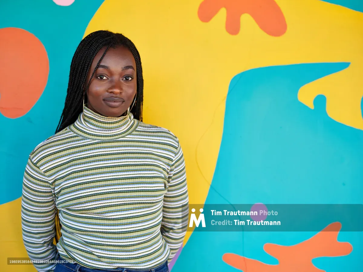 Naomi Likayi stands confidently before her completed mural in Portland's Alberta Arts District, wearing a striped turtleneck that echoes the rhythmic patterns of her artistic work. The explosive backdrop of turquoise, golden yellow, coral, and lavender organic shapes creates a dynamic canvas that transforms the Northeast Portland streetscape. Her serene expression and poised stance reflect the satisfaction of artistic completion, while the bold, flowing forms behind her pulse with contemporary energy against the urban setting.