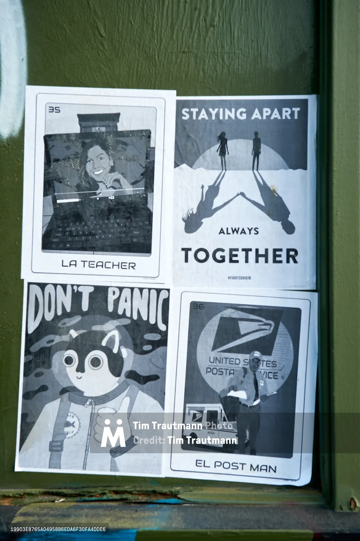 Four photocopied flyers create a makeshift gallery on weathered olive-green wood in Portland's Concordia neighborhood. The black and white prints capture pandemic zeitgeist through humor and hope: a smiling teacher from a Polaroid labeled "LA TEACHER," silhouettes maintaining distance under "STAYING APART ALWAYS TOGETHER," a cartoon astronaut proclaiming "DON'T PANIC," and "EL POST MAN" featuring a postal worker and truck. The grassroots display transforms ordinary street surface into community commentary during COVID-19 lockdown.
