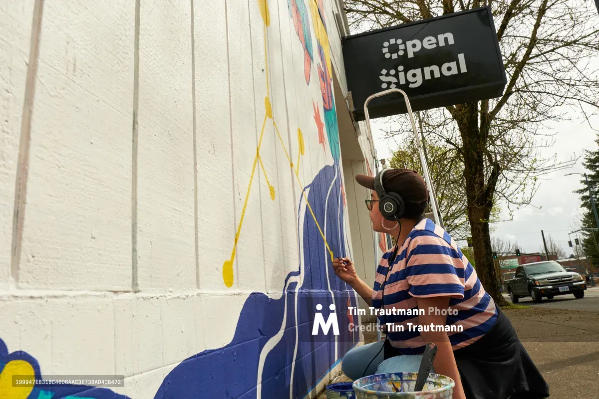 Maria Rodriguez, known as Sparkykneecap, methodically applies blue paint to her vibrant mural "Let's Talk" on the white exterior wall of Open Signal in Portland's Eliot neighborhood. Wearing wireless headphones and a striped shirt, the Mexican-American artist works with focused concentration as afternoon light illuminates her colorful work-in-progress. The scene captures the intimate process of public art creation, with the artist's paint bucket and brushes arranged nearby as she brings themes of identity and culture to life on Northeast Martin Luther King Jr. Boulevard.