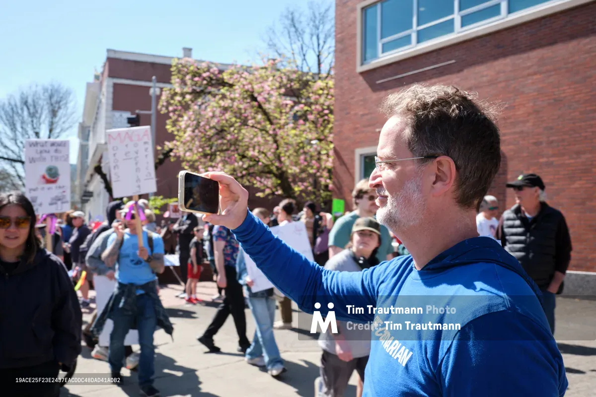 A middle-aged man in a blue hoodie raises his smartphone to document the scene at a protest march in Portland's historic Old Town district. Behind him, demonstrators carry signs and gather on Southwest Ankeny Street, while spring cherry blossoms frame the brick buildings. The bright afternoon light illuminates the crowd as the man, identified as Tim Dickinson, captures the moment of civic engagement unfolding around him.