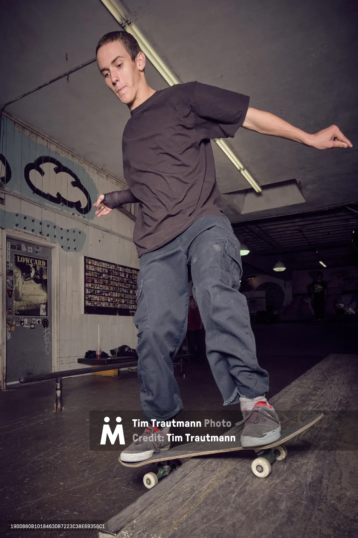 A focused skateboarder in muted earth tones navigates a weathered skateboard through the industrial interior of Commonwealth Skateboarding in Portland's Buckman neighborhood. Harsh fluorescent lighting cuts through the gritty atmosphere, casting stark shadows across concrete floors while vintage skate posters and graffiti-marked walls frame the scene. The rider's concentrated expression and fluid stance capture the meditative precision of street skating culture within this authentic underground retail space.