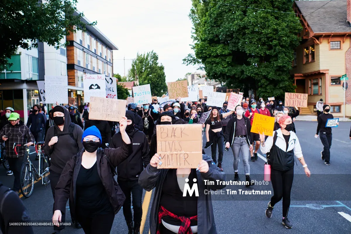 Masked protesters fill the asphalt streets of Portland's Kerns neighborhood during a Black Lives Matter demonstration in the aftermath of George Floyd's murder. The diverse crowd moves through the tree-lined residential area near East Burnside Street, carrying handmade signs with messages of justice and solidarity against a backdrop of characteristic Pacific Northwest architecture. The late afternoon light casts long shadows across the pavement as the peaceful march progresses through this historic Portland community.