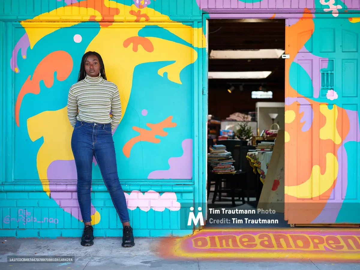 Naomi Likayi stands confidently against her newly completed mural in Portland's Alberta Arts District, where swirling abstract forms in electric turquoise, sunshine yellow, coral orange, and lavender purple transform the storefront facade. The young Black artist wears a cream striped turtleneck and dark jeans, her relaxed pose contrasting with the explosive energy of her artwork that flows around the entrance to what appears to be a community bookstore. Natural daylight illuminates both the artist and her bold street art, creating a powerful portrait of creative accomplishment in this gentrifying Northeast Portland neighborhood.
