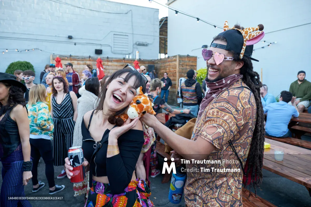 Two friends embrace the playful spirit of Your Sunday Best's season opener at White Owl Social Club, their laughter infectious as they sport novelty animal headwear and heart-shaped sunglasses. The woman in black clutches a plush fox while her companion grins beneath a giraffe headband, their joy radiating through the crowded rooftop venue. String lights overhead and the casual crowd create an atmosphere of communal celebration in this intimate Portland gathering.