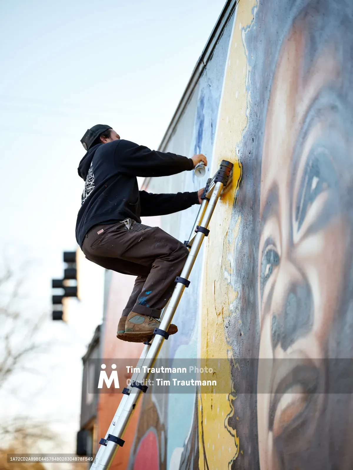 Travis Fields, known as campographic, balances on a telescoping ladder against a weathered wall, applying vibrant yellow paint to his Amanda Gorman mural at Northeast 15th and Killingsworth in Portland's Alberta district. The artist works with focused concentration under overcast skies, his dark hoodie and beanie contrasting against the emerging colors of the tribute piece. Urban residential buildings blur softly in the background, creating depth while emphasizing the intimate scale of this community art project.