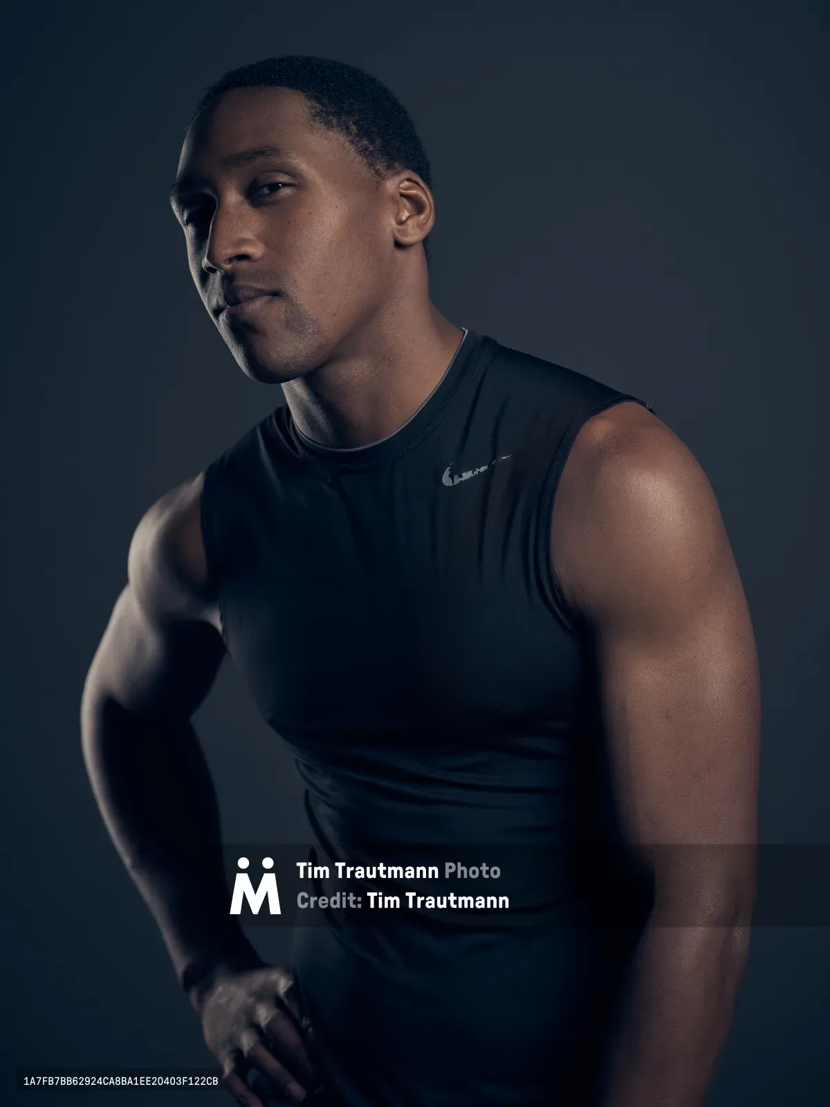 A contemplative athletic portrait captured during the Fujifilm GFX 100 launch tour in Portland's Northwest District. The subject, a young Black man in his twenties, gazes thoughtfully to the side while wearing a sleeveless black athletic shirt. Dramatic chiaroscuro lighting sculpts his muscular form against a deep charcoal backdrop, creating a powerful study in strength and introspection. The precise lighting emphasizes the definition of his shoulders and arms while casting compelling shadows across his face.