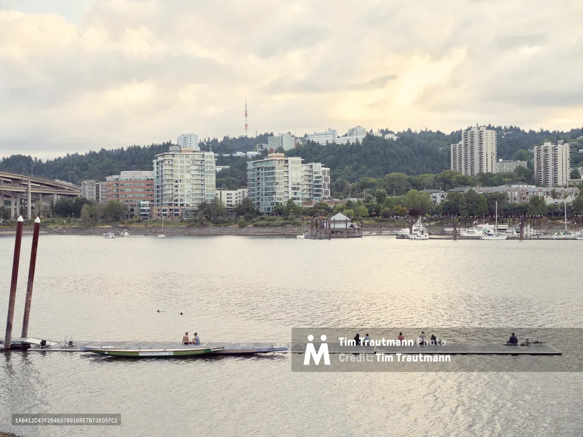 People gather along the peaceful waters of the Willamette River during early evening at Holman Dock, enjoying the tranquil waterfront setting. The towering medical complex of Oregon Health and Science University rises from the forested hillside beyond, its glass facades catching the muted light beneath overcast skies. The serene marina setting frames this moment of leisure, where urban development meets recreational relaxation along Portland's waterfront.