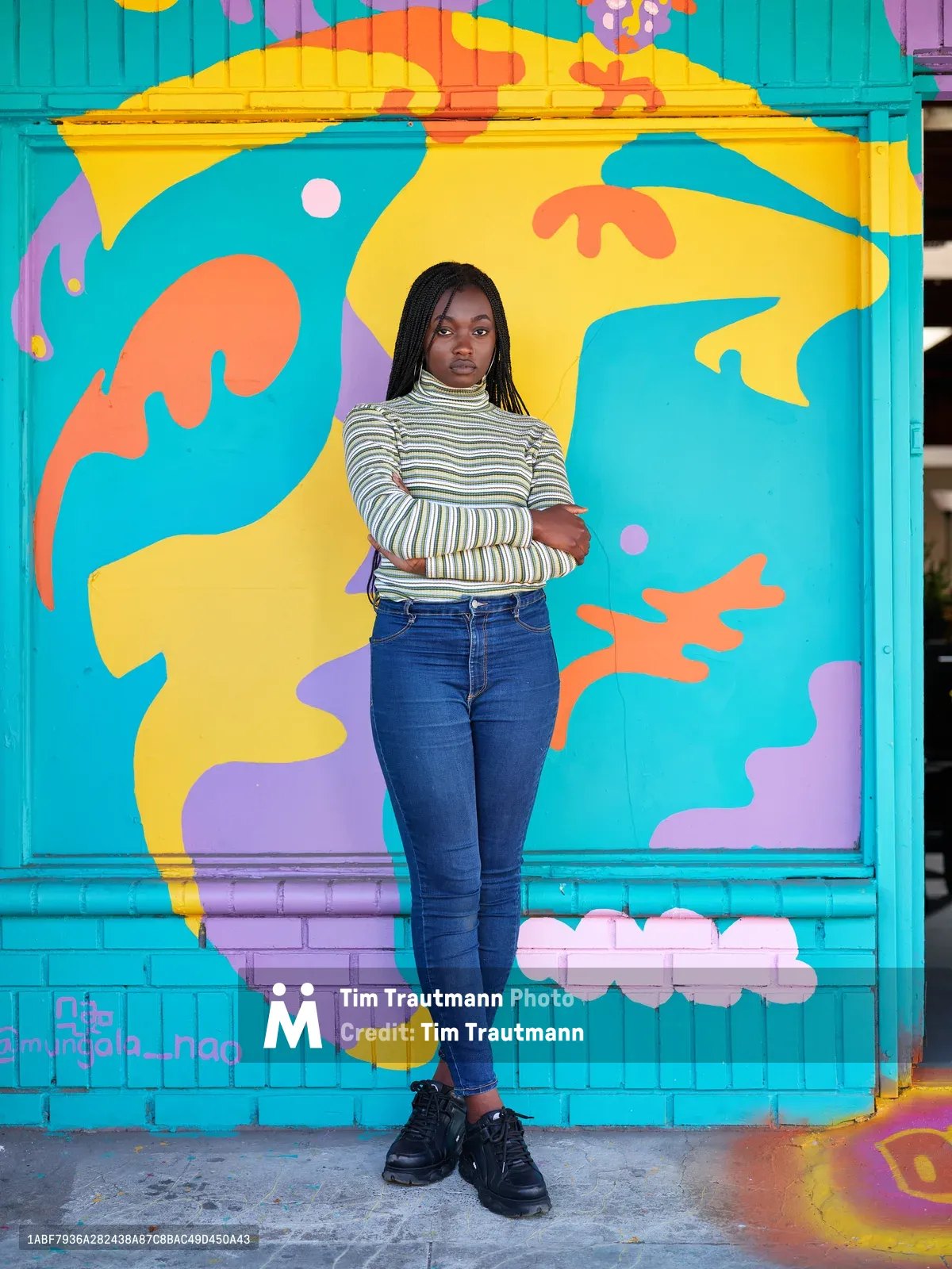 Naomi Likayi stands confidently with crossed arms before her completed mural in Portland's Alberta Arts District. The artist wears a striped turtleneck and dark jeans, positioned against an explosion of turquoise, yellow, orange, and purple abstract forms that dance across the brick wall behind her. Natural daylight illuminates both subject and artwork, creating a harmonious dialogue between creator and creation in this Northeast Portland neighborhood known for its thriving street art scene.