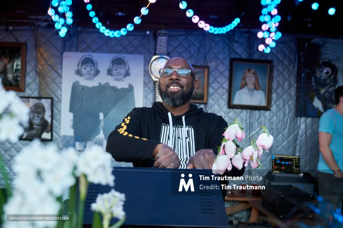 DJ Cee White, wearing reflective sunglasses and a black hooded sweatshirt, commands the decks at White Owl Social Club's intimate venue space. Cascading string lights create ethereal bokeh patterns above vintage portrait photographs that line the diamond-quilted walls, while pink tulips in the foreground add unexpected softness to this Portland nightlife scene. The atmospheric lighting and eclectic gallery setting transform the day party into a moody sonic sanctuary.
