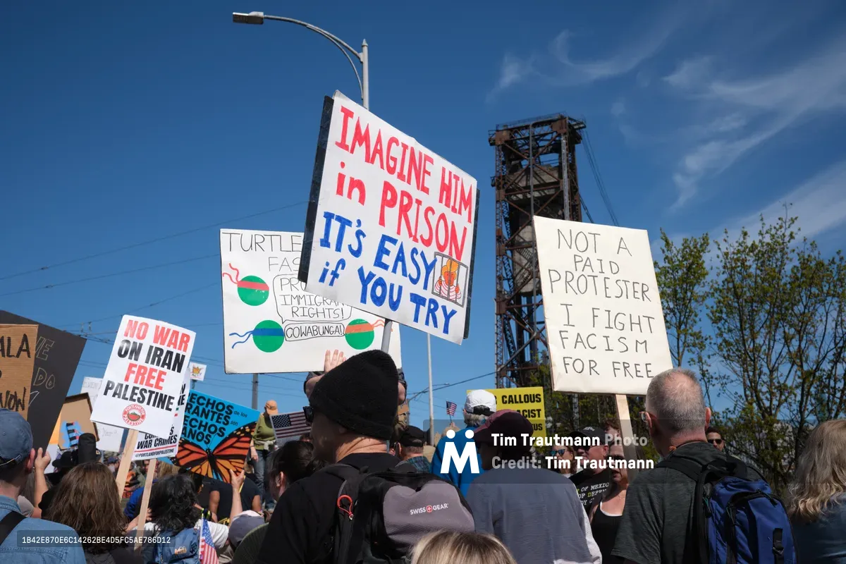 Under a brilliant cobalt sky, protesters gather on Portland's Steel Bridge holding handmade signs with bold messages including "IMAGINE HIM IN PRISON" and "NOT A PAID PROTESTER I FIGHT FACISM FOR FREE." The crowd flows beneath an iconic steel truss structure, with the industrial bridge architecture framing this grassroots demonstration. Diverse participants of varying ages clutch their signs aloft, their faces partially obscured but their collective energy palpable in the crisp daylight.