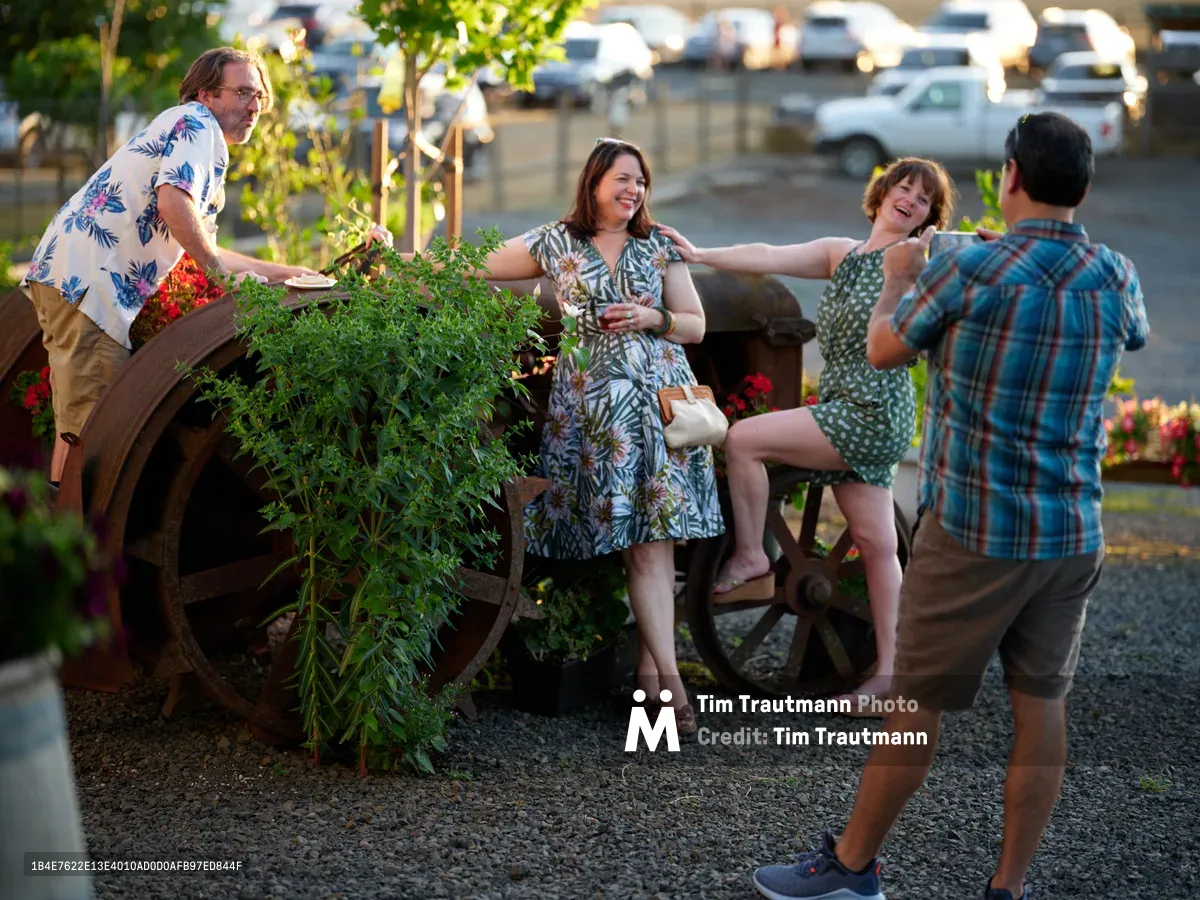Four friends share an intimate moment of connection around a rustic wooden cart overflowing with fresh herbs and vibrant flowers at Abbey Road Farm in Carlton, Oregon. The warm golden hour light filters through the scene as two women sit casually on the cart while a bearded man in a tropical print shirt tends to the plants and another friend captures the moment. The organic farm setting creates an atmosphere of authentic community and agricultural abundance, with vehicles softly blurred in the background suggesting the accessible yet pastoral nature of this Willamette Valley location.