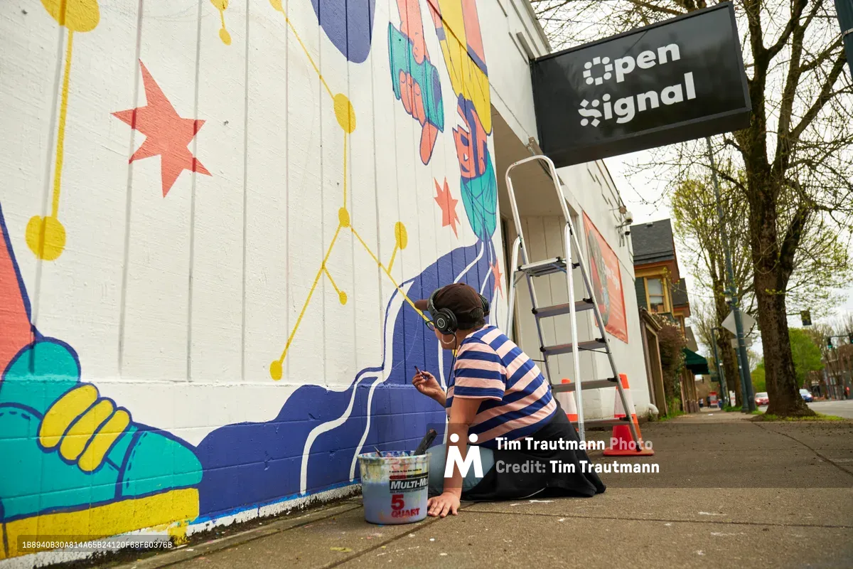 Mexican-American artist Maria Rodriguez, known as Sparkykneecap, kneels on the sidewalk while adding vibrant blue details to her mural "Let's Talk" on the white exterior wall of Open Signal in Portland's Eliot neighborhood. The large-scale artwork features playful geometric shapes, whimsical characters, and bold colors including coral stars and turquoise elements that reflect themes of identity and cultural connection. A stepladder and paint supplies rest nearby as the artist works under the filtered light of an overcast day, with the Open Signal sign visible above and bare spring trees framing the urban scene.