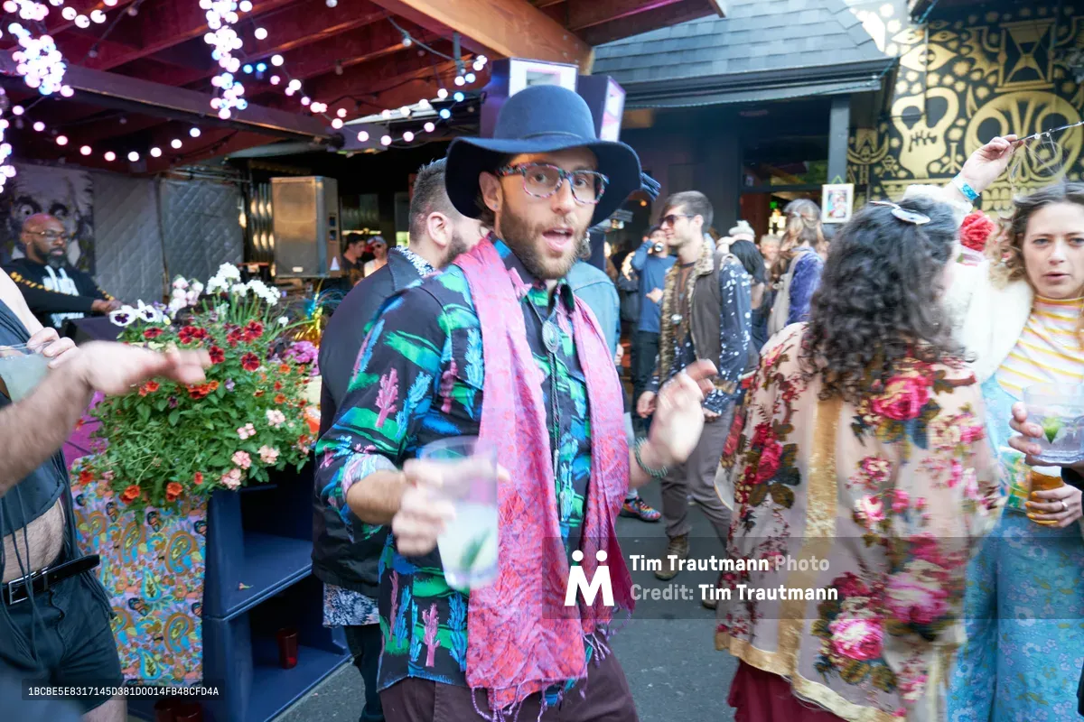 A vibrant scene unfolds at White Owl Social Club as a bearded man in a wide-brimmed hat and kaleidoscopic floral shirt gestures animatedly amid the bustling Your Sunday Best day party. String lights twinkle overhead while colorful flowers and urban murals create a bohemian backdrop, capturing the eclectic energy of Portland's summer social scene. The motion blur of party-goers and warm lighting convey the infectious atmosphere of this popular seasonal gathering.