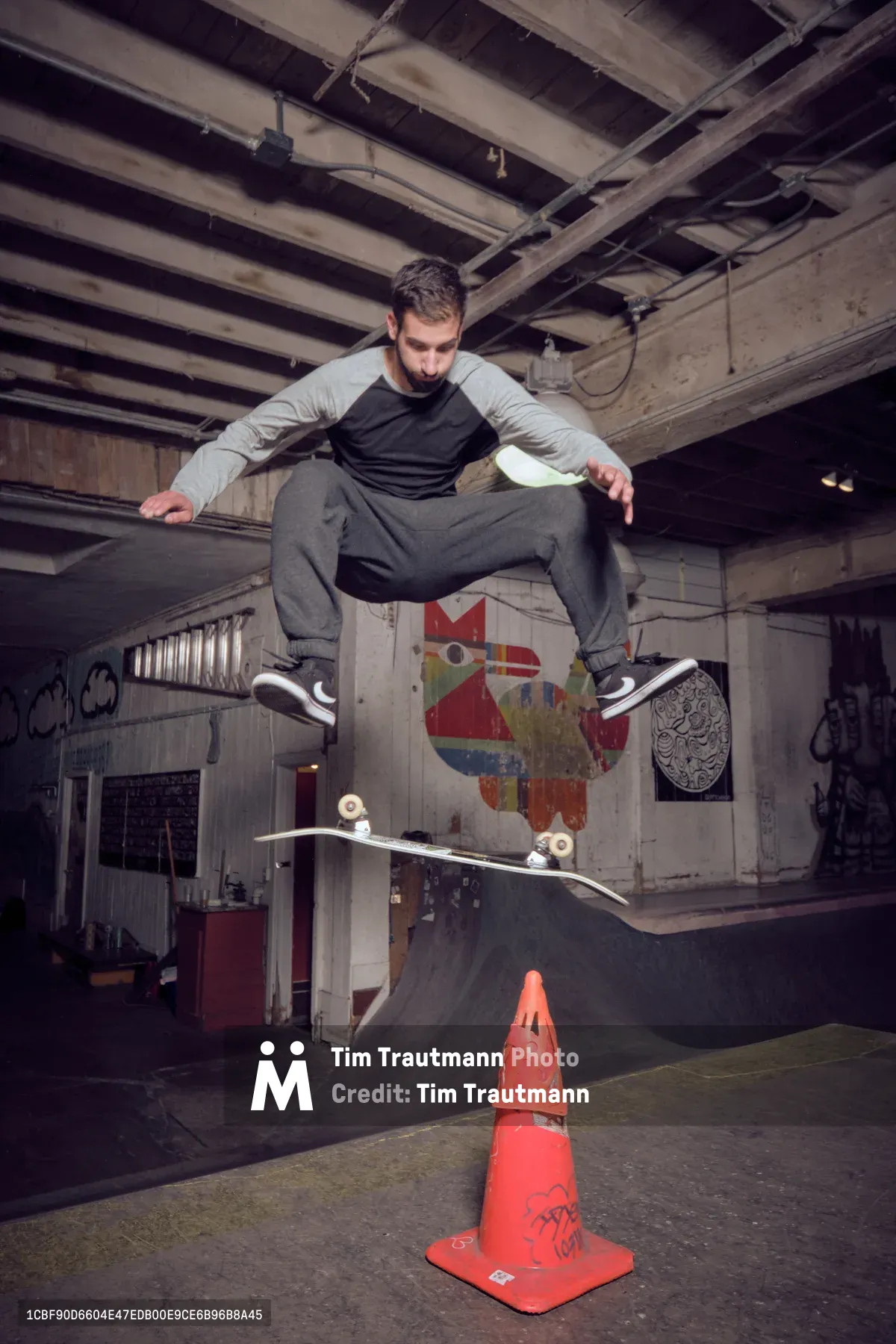 A focused skateboarder executes a precise ollie over a weathered orange traffic cone in the atmospheric basement of Commonwealth Skateboarding in Portland's Buckman neighborhood. The industrial space features exposed ceiling joists, graffitied concrete walls, and a makeshift skate bowl, while warm overhead lighting illuminates the concentrated athlete mid-trick. The gritty urban interior, with its painted murals and raw concrete surfaces, captures the authentic underground skateboarding culture of Southeast Portland.