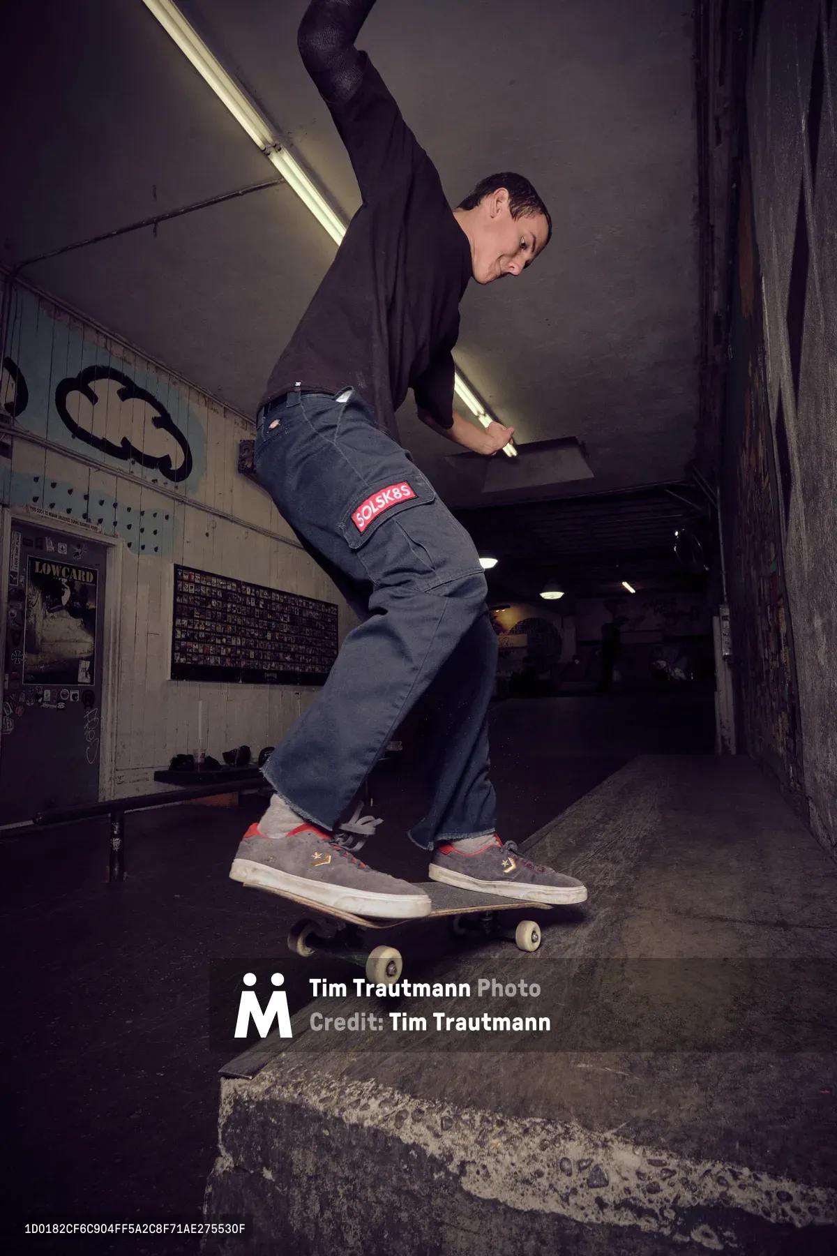 A focused skateboarder in gray work pants and casual sneakers glides through the interior of Commonwealth Skateboarding in Portland's Buckman neighborhood. The atmospheric scene captures him mid-ride beneath harsh fluorescent lighting, with vintage skateboard graphics and weathered concrete steps creating an authentic underground skate culture backdrop. His concentrated expression and fluid motion through this iconic Portland skate shop epitomize the dedication and flow that defines street skating.