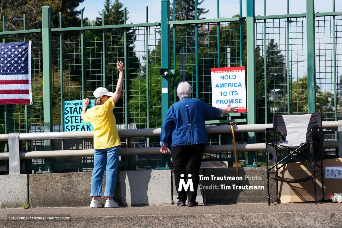 Two protesters position themselves along the North Skidmore overpass above Interstate 5 in Portland's Humboldt neighborhood, displaying signs calling for civic engagement and a May 1st General Strike. A woman in a bright yellow shirt raises her hand in greeting while an elderly man with a walking cane adjusts a red, white and blue protest banner reading 'Hold America to Its Promise.' The green mesh security fencing and concrete barriers frame their grassroots demonstration against the backdrop of evergreen trees under overcast Pacific Northwest skies.