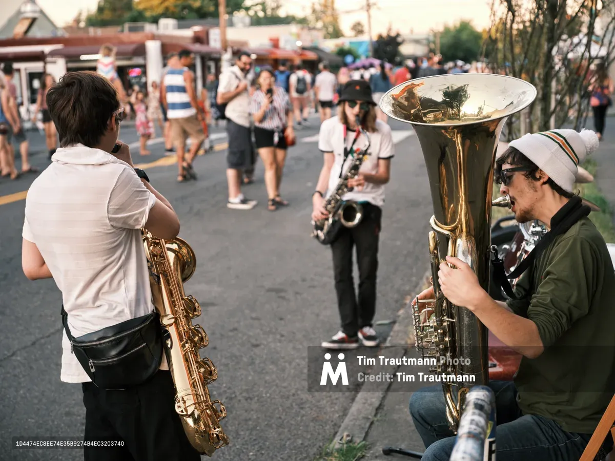 Three street musicians create an impromptu brass ensemble on Northeast Alberta Street during Portland's Last Thursday art walk. A saxophonist in a white hoodie stands alongside a seated tuba player wearing a knit beanie, while a third musician with a bass clarinet completes their urban trio. The golden hour light bathes the scene as festival-goers drift past the shuttered storefronts and food trucks in the background, capturing the authentic spirit of this monthly celebration that transforms Alberta Street into a pedestrian playground.