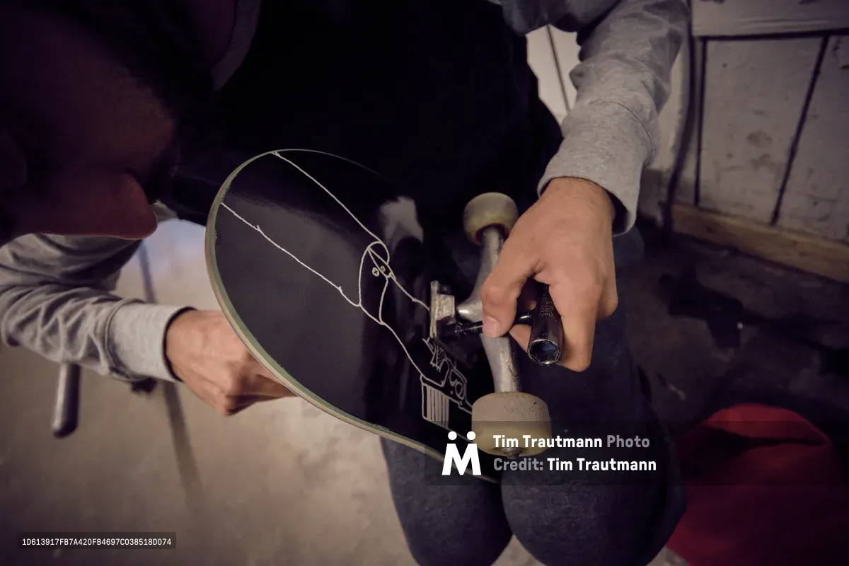 In the intimate workspace of Commonwealth Skateboarding on Southeast 20th Avenue, focused hands adjust the trucks and wheels of a black skateboard deck adorned with linear graphics. The moody lighting casts deep shadows across weathered concrete floors, while the skater's deliberate movements speak to the meticulous craft of board assembly. This behind-the-scenes moment captures the tactile relationship between rider and equipment in Portland's vibrant skate culture.