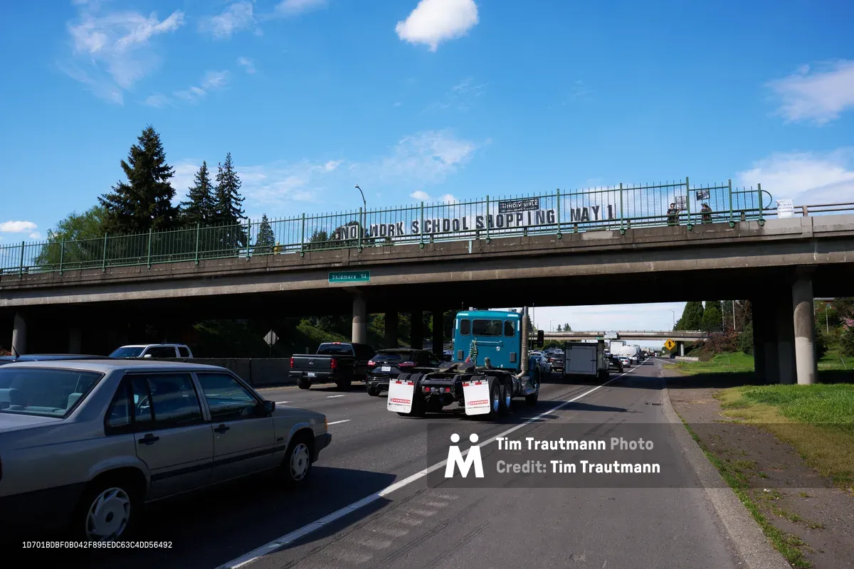 Activists from the Indivisible movement unfurl a protest banner reading 'No Work School Shopping May 1st' across the North Skidmore Street overpass above Interstate 5 in Portland's Humboldt neighborhood. Heavy afternoon traffic flows beneath the concrete bridge as towering evergreens frame the scene under partly cloudy Pacific Northwest skies. The grassroots demonstration calls for a general strike, transforming urban infrastructure into a platform for political messaging.