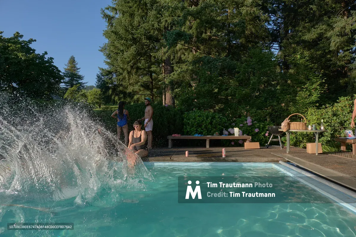 A swimmer creates a dramatic splash as they dive into the turquoise waters of a backyard pool nestled within the lush greenery of Newberg's Orchard Vibration. The scene captures a perfect summer afternoon with friends gathered around the pool deck, surrounded by towering evergreens and vibrant foliage that creates an intimate, secluded atmosphere. Golden hour light filters through the canopy, illuminating the crystalline water and the rustic wooden deck furniture that speaks to Oregon's casual outdoor living culture.
