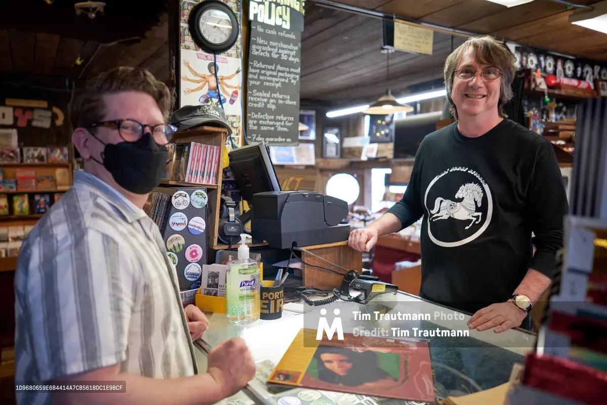 Two music enthusiasts connect across the glass counter at Music Millennium during Record Store Day 2022, their shared passion for vinyl evident in the warm exchange. The masked customer in plaid shirt leans forward while the店owner in black long-sleeve shirt with circular logo stands behind the register, surrounded by the eclectic chaos of badges, policy signs, and musical memorabilia that defines this iconic Portland record shop. Fluorescent lighting bathes the scene in the authentic glow of independent retail culture, capturing a moment where analog music brings people together.
