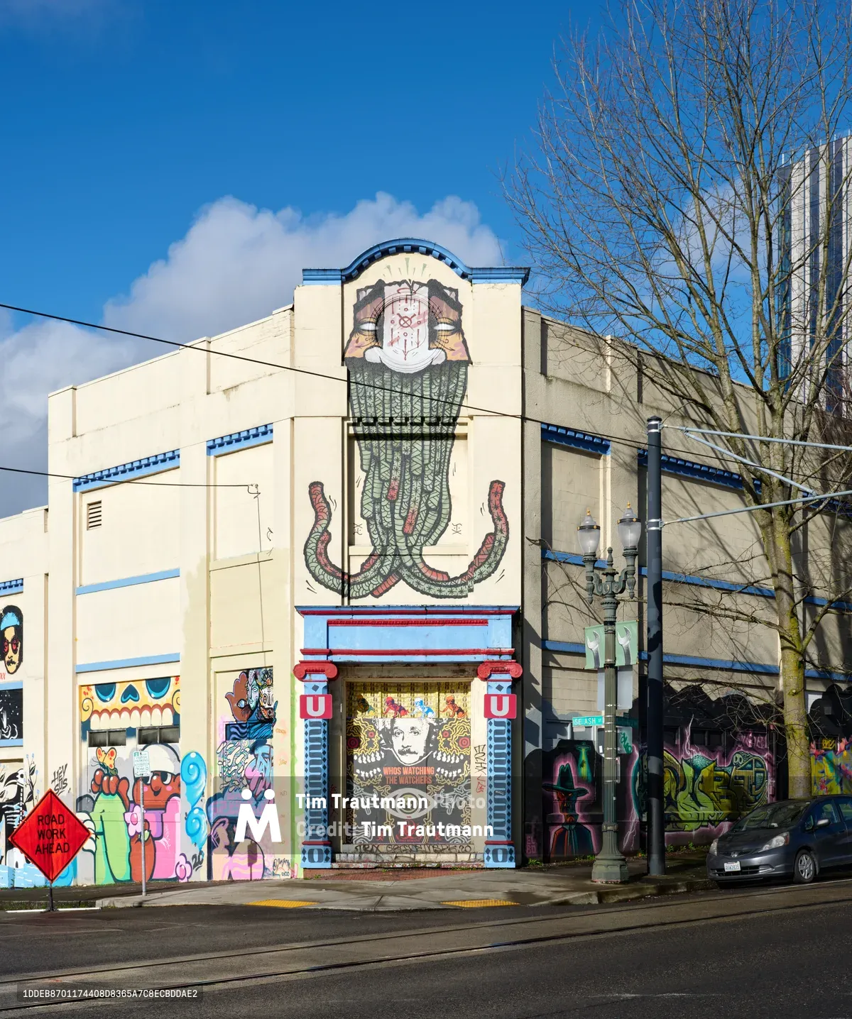 A towering serpentine deity dominates the facade of a former Salvation Army warehouse in Portland's Central Eastside district, its coiled form rising majestically between red classical columns. The intricate street mural features a crowned figure with an all-seeing eye, rendered in earth tones against the building's cream-colored walls under brilliant blue winter skies. Vibrant graffiti art adorns the structure's lower sections, while bare tree branches frame the composition and power lines cross the urban canvas.