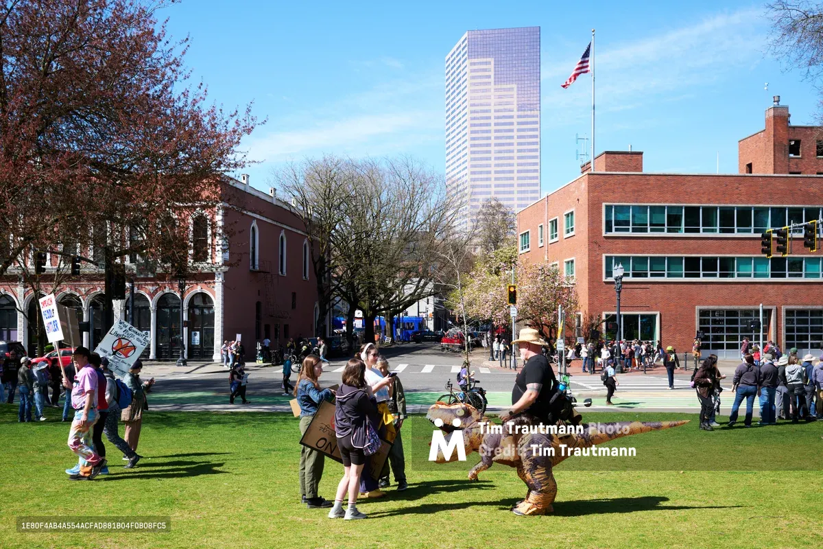 Protesters gather in Portland's historic Skidmore Fountain area during a 'No Kings' demonstration, with participants in inflatable dinosaur costumes adding whimsical defiance to the political assembly. The scene unfolds beneath clear spring skies, framed by the district's characteristic red brick architecture and a gleaming downtown tower. Families with children mingle among activists holding signs, creating an atmosphere where civic engagement meets carnival spirit in the heart of Old Town.