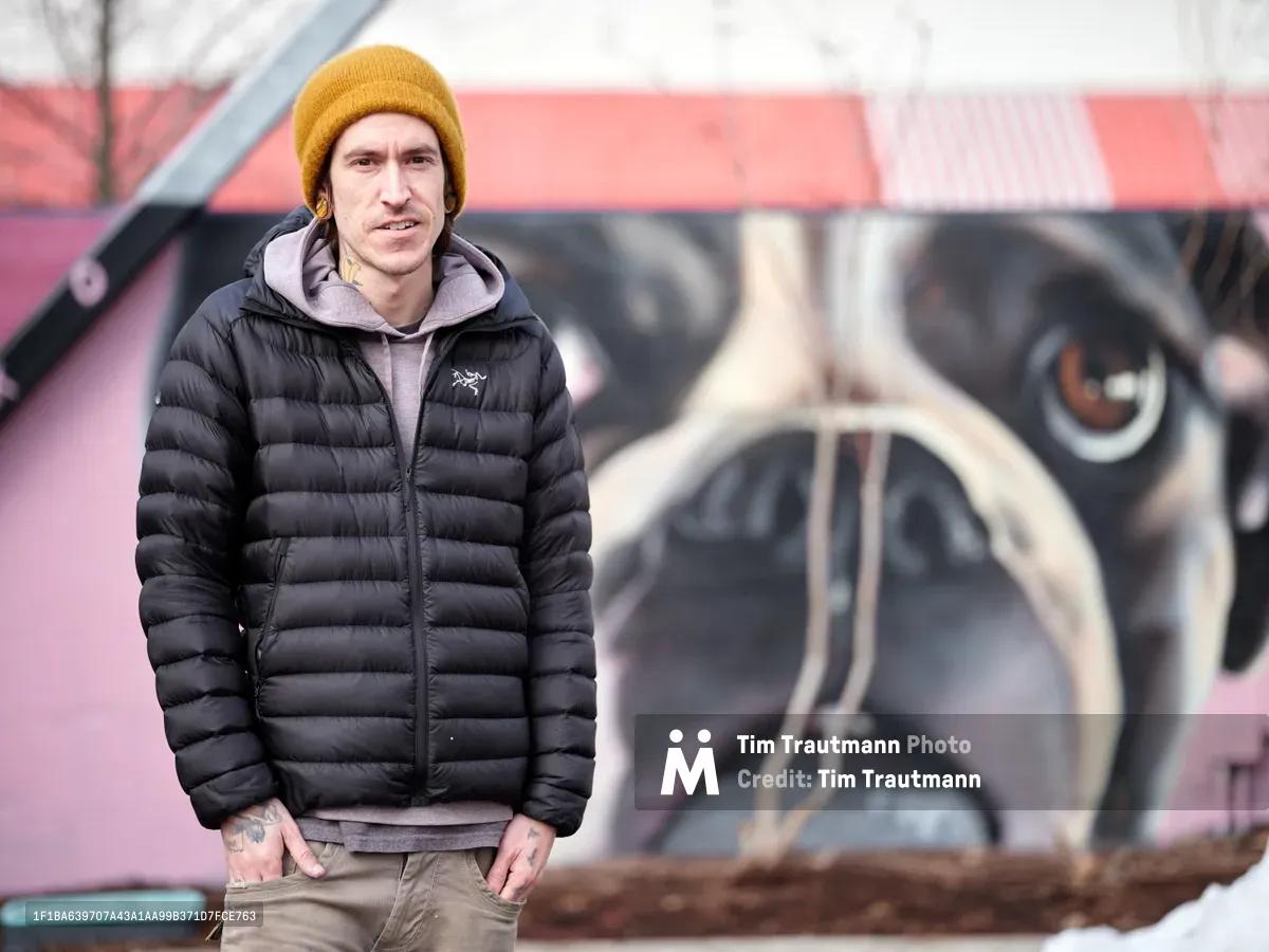 Muralist Weti stands confidently before his vibrant street art creation on the exterior wall of Clay Creative Building in Southeast Portland's Hosford-Abernethy neighborhood. Wrapped in a black down jacket and golden knit beanie against the Pacific Northwest chill, the tattooed artist poses with quiet satisfaction beside his large-scale work featuring bold geometric forms and watchful eyes rendered in pink, black, and cream tones. The urban portrait captures both the creator and creation in sharp focus, while the colorful mural provides a dynamic backdrop that speaks to Portland's thriving street art culture.