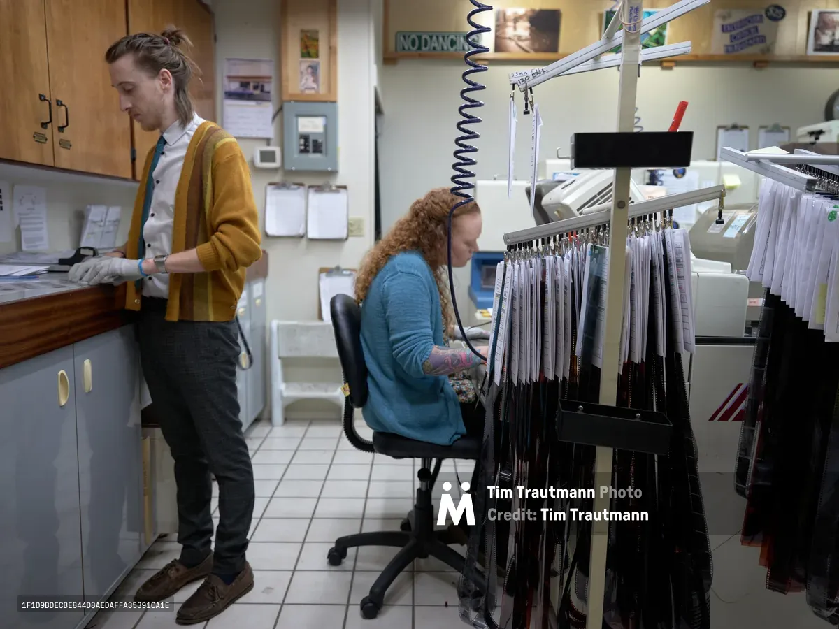 In the intimate workspace of Blue Moon Camera & Machine on North Lombard Street, two technicians pursue the meticulous craft of analog photography development. A young man in a golden cardigan examines film strips at a light table while his colleague, wearing a blue sweater, operates specialized processing equipment surrounded by hanging negatives. The fluorescent-lit lab, dense with filing systems and vintage photography notices, captures the enduring artistry of traditional film processing in Portland's Saint Johns neighborhood.