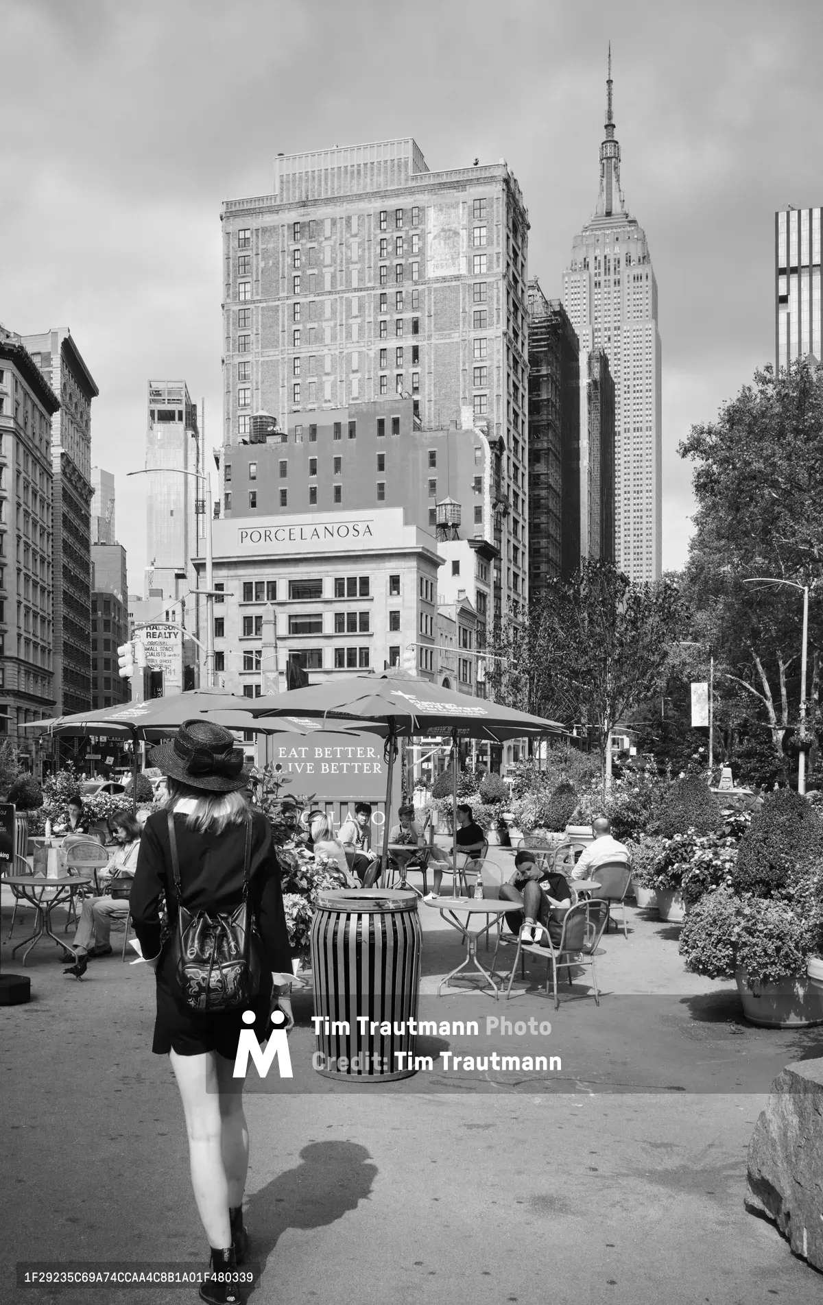 A contemplative woman in a wide-brimmed hat pauses at the edge of Madison Square Park's bustling outdoor dining area, her shadow stretching across sun-warmed concrete. The Empire State Building rises majestically in the background, its Art Deco spire piercing overcast skies, while the historic Porcelanosa building and contemporary high-rises create Manhattan's signature architectural layering. The scene captures the gentle rhythm of urban life, where nature and commerce intersect in temporary harmony beneath market umbrellas and flowering planters.