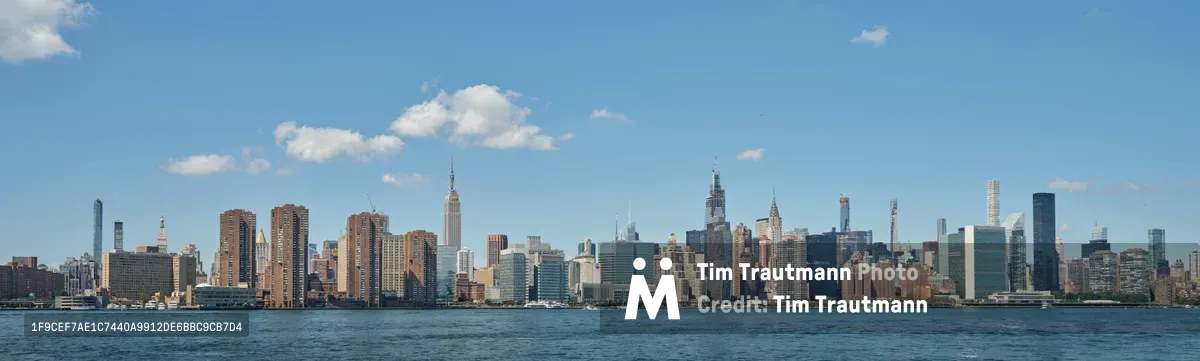 The iconic Manhattan skyline rises majestically across the East River, viewed from the industrial waterfront of Greenpoint, Brooklyn. Wispy white clouds drift across an azure afternoon sky, creating a serene backdrop for the dense cluster of skyscrapers including the distinctive Empire State Building. The foreground features weathered wooden pier pilings emerging from the dark river waters, adding rustic texture to this classic New York vista. The composition captures the dramatic contrast between Brooklyn's quieter shores and Manhattan's towering urban density.