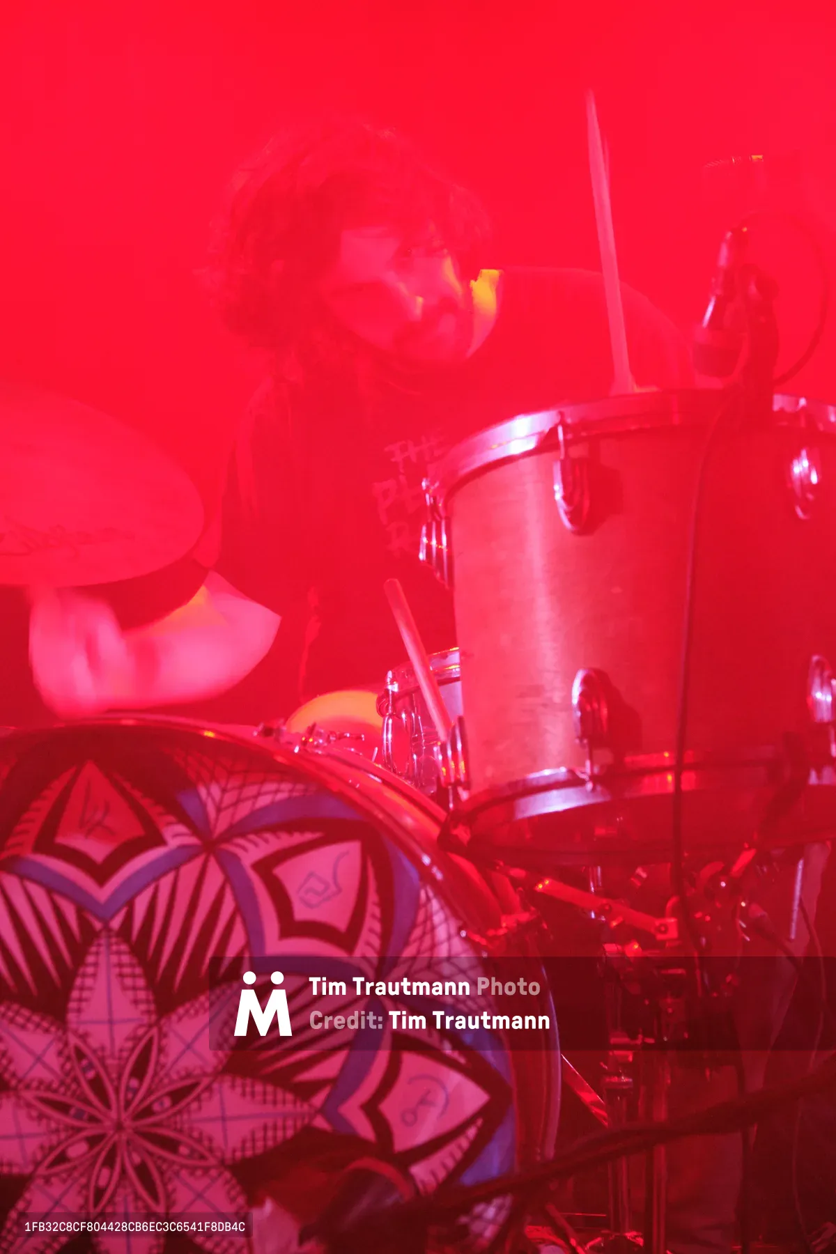 A drummer from experimental band Sun Blood Stories performs behind a drum kit awash in deep red stage lighting at Dante's venue in downtown Portland. The intense crimson wash creates an ethereal, almost otherworldly atmosphere, transforming the geometric patterns on the drum kit into shadowy mandala-like designs. Steam or haze drifts through the saturated light, while the musician remains a silhouetted figure within this scarlet-soaked sonic landscape.