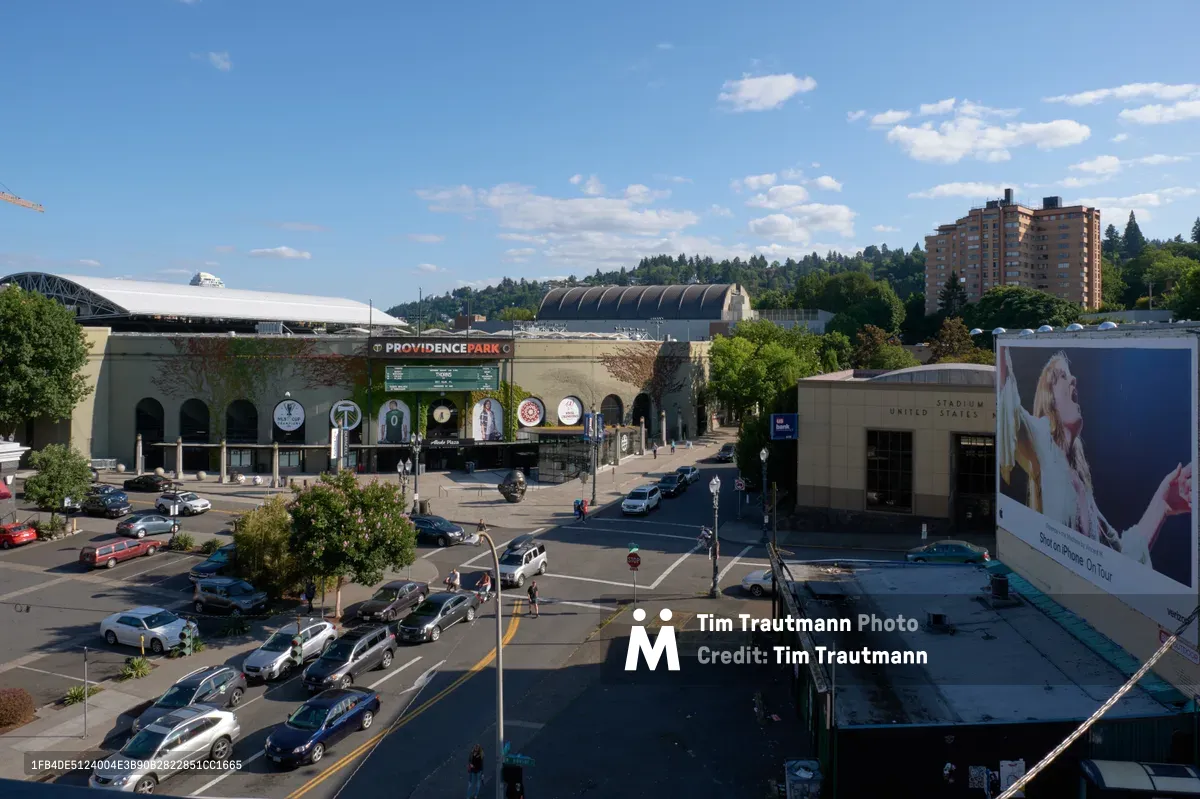 An elevated view captures the bustling intersection surrounding Providence Park in Portland's Goose Hollow neighborhood on a clear afternoon. The iconic soccer stadium anchors the frame while residential towers rise against forested West Hills in the distance. Street-level activity flows around the venue as cars navigate the urban intersection, with commercial signage and modern architecture defining this vibrant sports district.