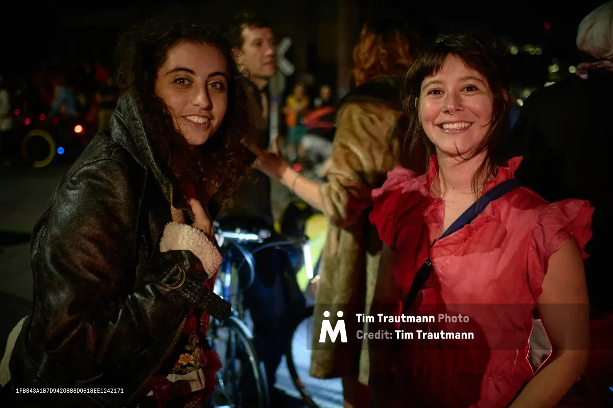Two young women beam with infectious joy during Portland's Drop Out Prom Ride, their contrasting outfits—one in weathered olive military jacket, another in vibrant coral ruffled dress—creating a striking visual dialogue against the blurred nocturnal revelry. The warm glow of streetlights and scattered bicycle lights illuminate their genuine smiles, capturing the spontaneous intimacy of friendship amid the organized chaos of Pedalpalooza's unconventional celebration in Southeast Portland's Hosford-Abernethy neighborhood. Behind them, fellow participants blur into impressionistic figures, their presence suggesting the communal energy of this beloved alternative prom experience.