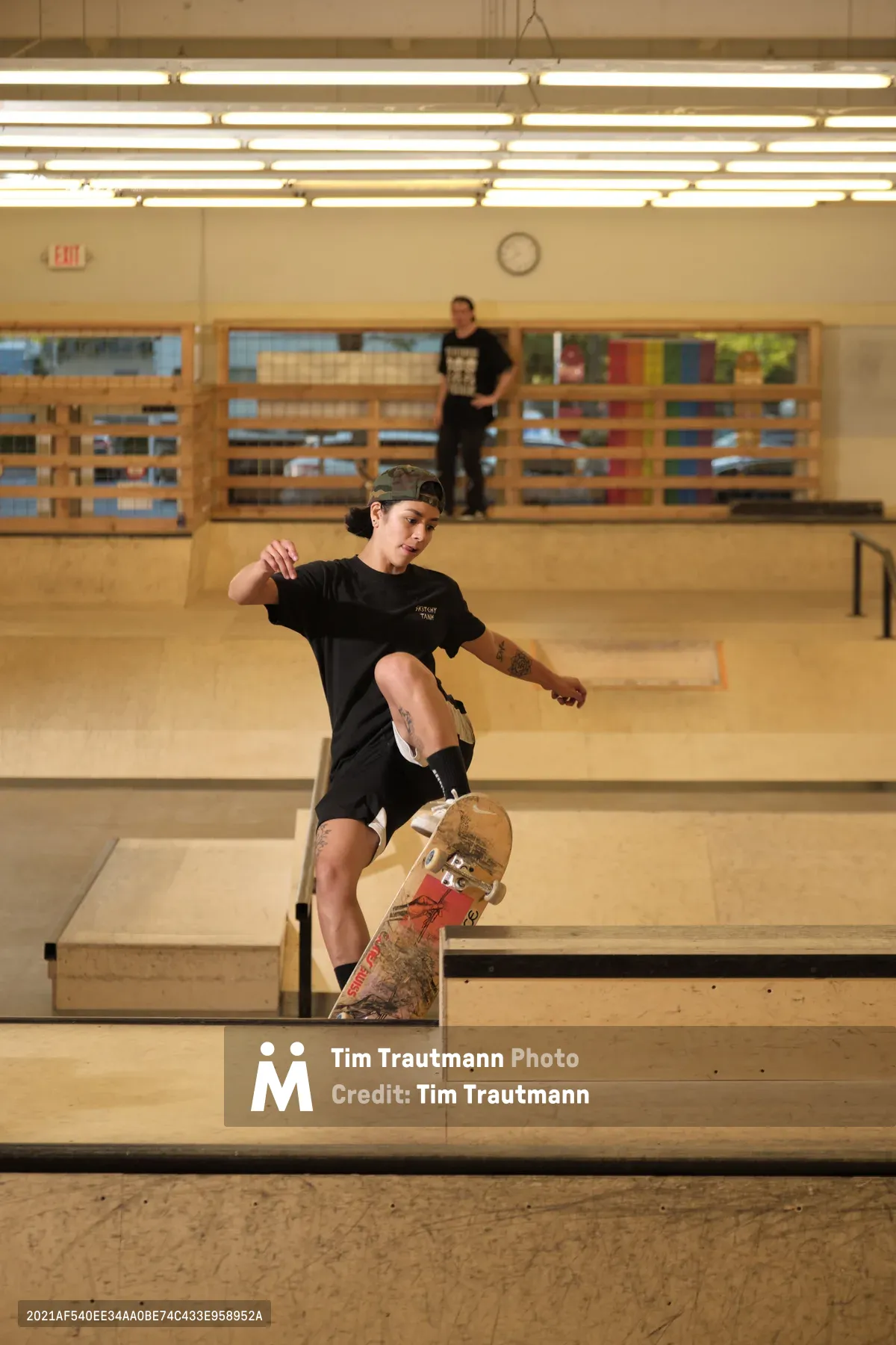 Professional skateboarder Christiana Means demonstrates fluid skateboarding technique as she navigates the curved concrete bowl at Stronger Skatepark in Milwaukie, Oregon. Her focused expression and athletic form are captured mid-maneuver, with warm fluorescent lighting illuminating the indoor facility's wooden observation deck and neutral concrete surfaces. A fellow skater observes from the elevated platform, creating depth in this dynamic action portrait.