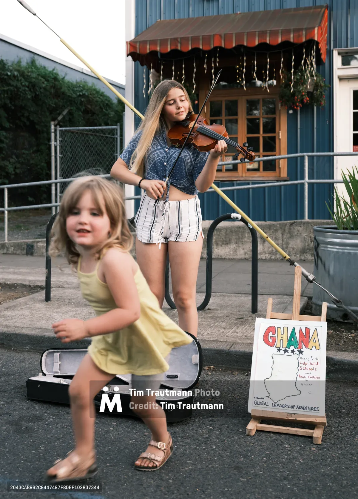 A teenage street musician plays violin with closed eyes and focused expression during Portland's Last Thursday street fair on Alberta Street. Her blonde hair catches the warm evening light as she performs in a denim top and striped shorts beside her open violin case. A young girl in a yellow dress dances joyfully in the foreground, embodying the spontaneous magic of community street performance. Behind them, a colorful sign promoting Ghana educational programs adds cultural context to this vibrant neighborhood gathering.