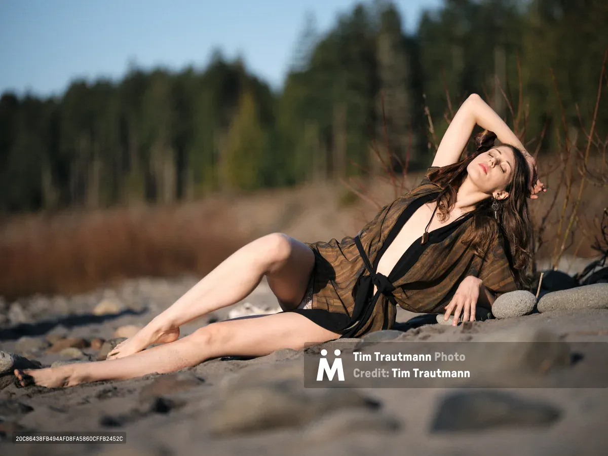 A brunette woman in a flowing black bikini and sheer cover-up reclines gracefully on sandy riverbed, her eyes closed in peaceful contemplation. The warm golden light of late afternoon bathes the scene as she rests against tall marsh grasses, with the blurred forest of Oregon's Floodplain Trail creating a dreamy backdrop. Her relaxed pose, with one arm behind her head, captures a moment of pure tranquility in this natural riverside setting.