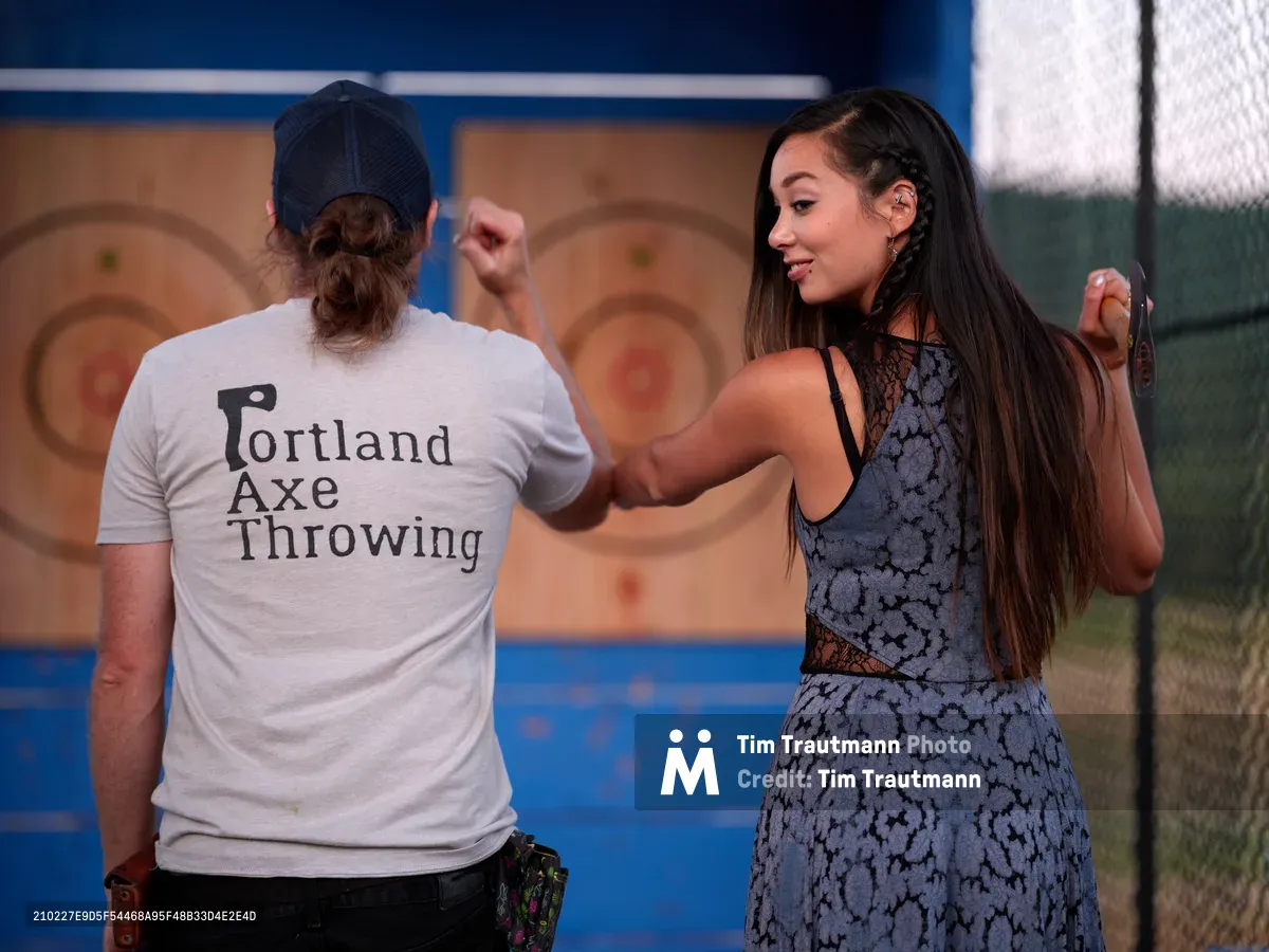 An instructor in a blue floral dress demonstrates proper axe throwing technique to a student wearing a Portland Axe Throwing t-shirt at Abbey Road Farm in Carlton, Oregon. The scene captures a moment of focused instruction against wooden target boards, with warm afternoon light filtering through the outdoor venue. The instructor's patient guidance contrasts with the student's concentrated posture as they prepare to release their throw.