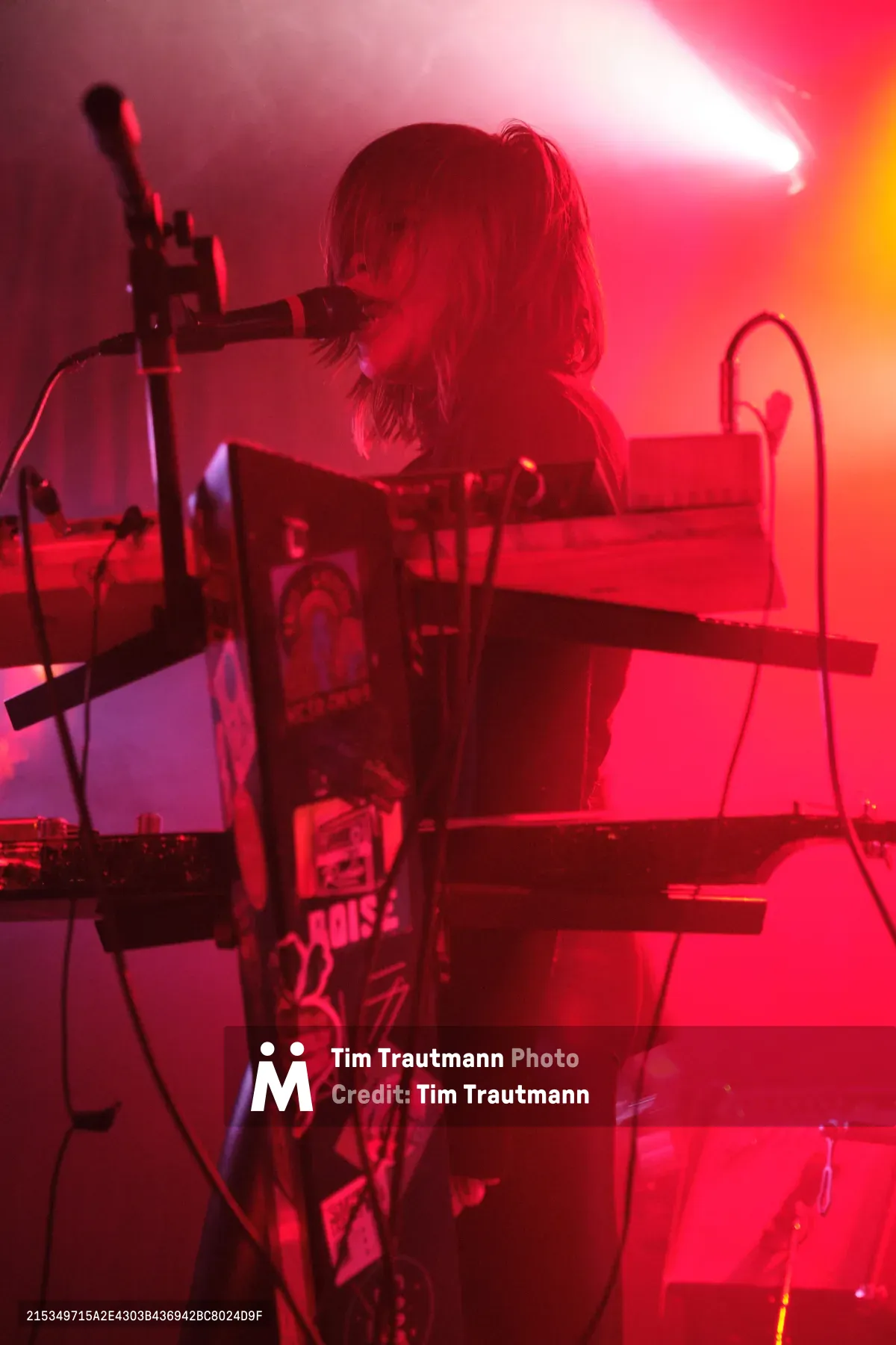 A keyboardist from Sun Blood Stories becomes silhouetted against a dramatic wash of crimson and magenta stage lighting at Dante's in downtown Portland. The performer's blonde hair catches the ethereal glow while they lean into their instrument, surrounded by synthesizer equipment and microphone stands. The atmospheric lighting transforms the intimate venue into a dreamlike sonic landscape, with deep reds bleeding into purple shadows.