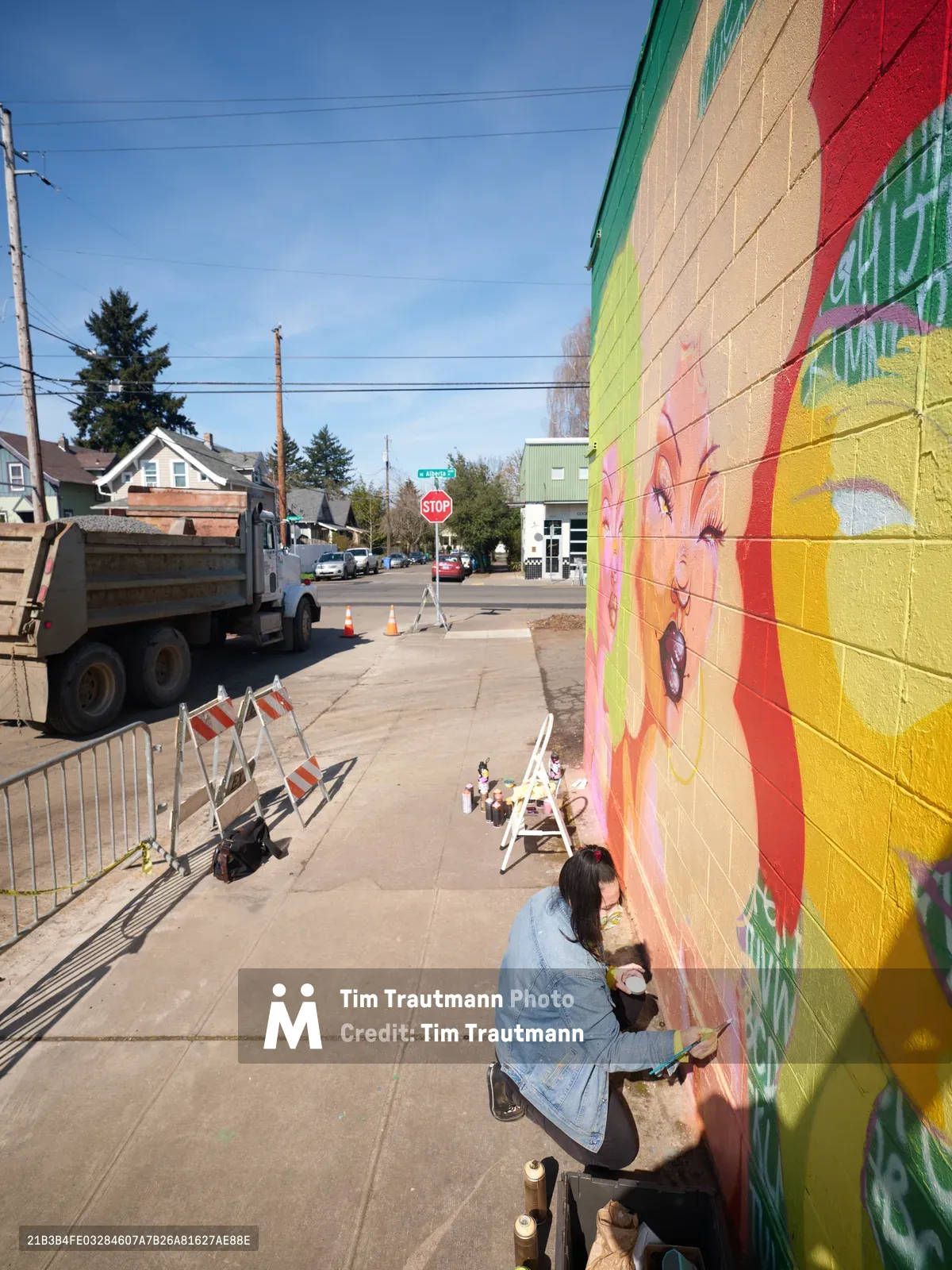Multidisciplinary artist Vilatose kneels on the sidewalk, adding delicate details to the luminous Jardín Cubana mural on Alberta Street in Portland's renowned arts district. The vibrant yellow and coral female figures emerge from blocks of masonry beneath crystalline blue skies, while construction barriers and urban infrastructure frame this moment of creative transformation. Paint containers scattered nearby capture the intimate process of public art creation in one of the Pacific Northwest's most culturally vibrant neighborhoods.
