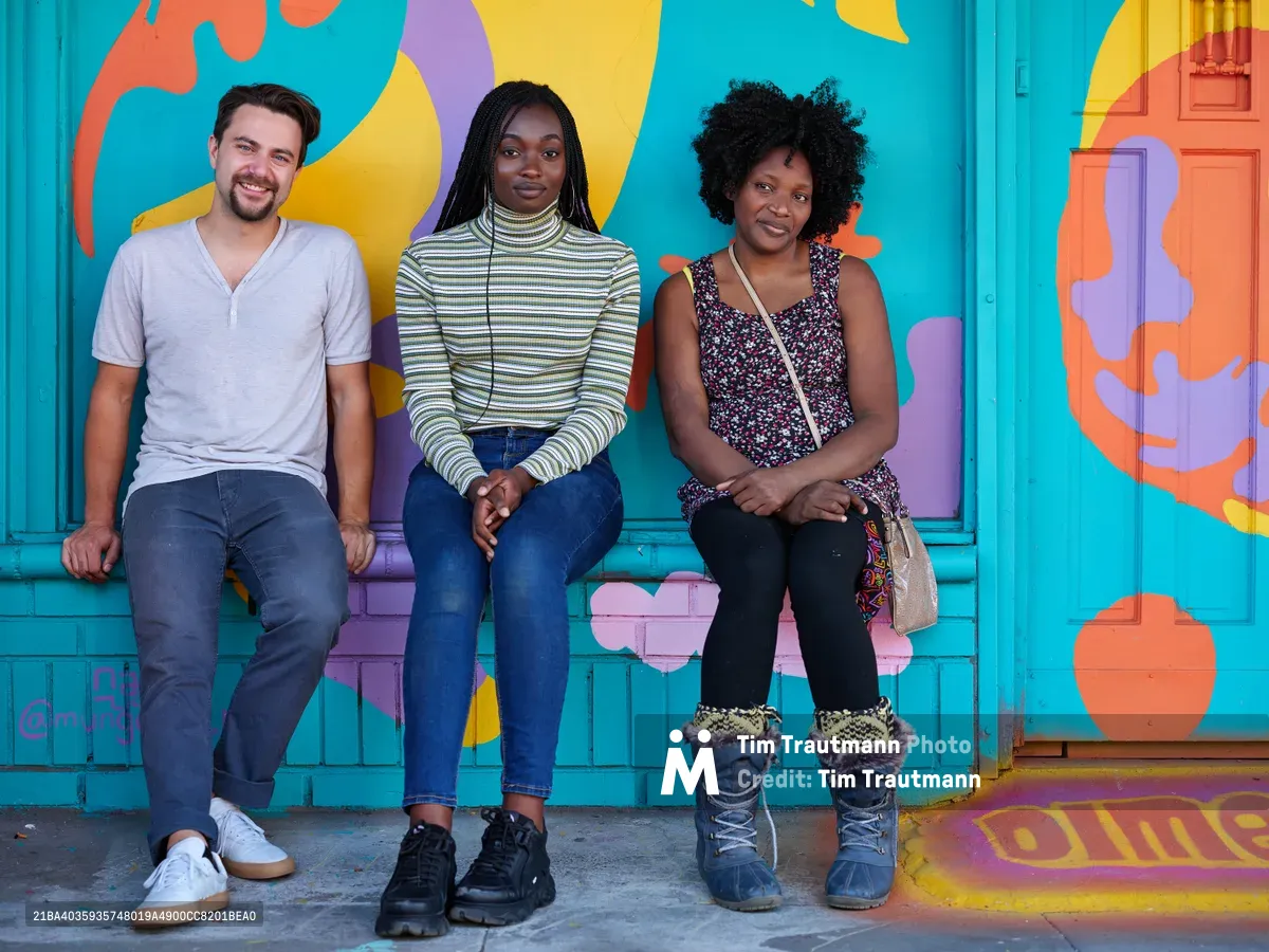 Travis Fields, Naomi Likayi, and the owner of the Dime & Penny sit together on the steps of a vibrant turquoise building adorned with bold abstract murals in Portland's Alberta Arts District. The trio radiates warmth and community spirit against the kaleidoscope of coral, yellow, and purple paint that transforms the urban storefront into a canvas of creative expression. Natural daylight illuminates their relaxed poses and genuine smiles, capturing an intimate moment of friendship and local business camaraderie. The colorful street art backdrop perfectly embodies the artistic energy that defines this Northeast Portland neighborhood.