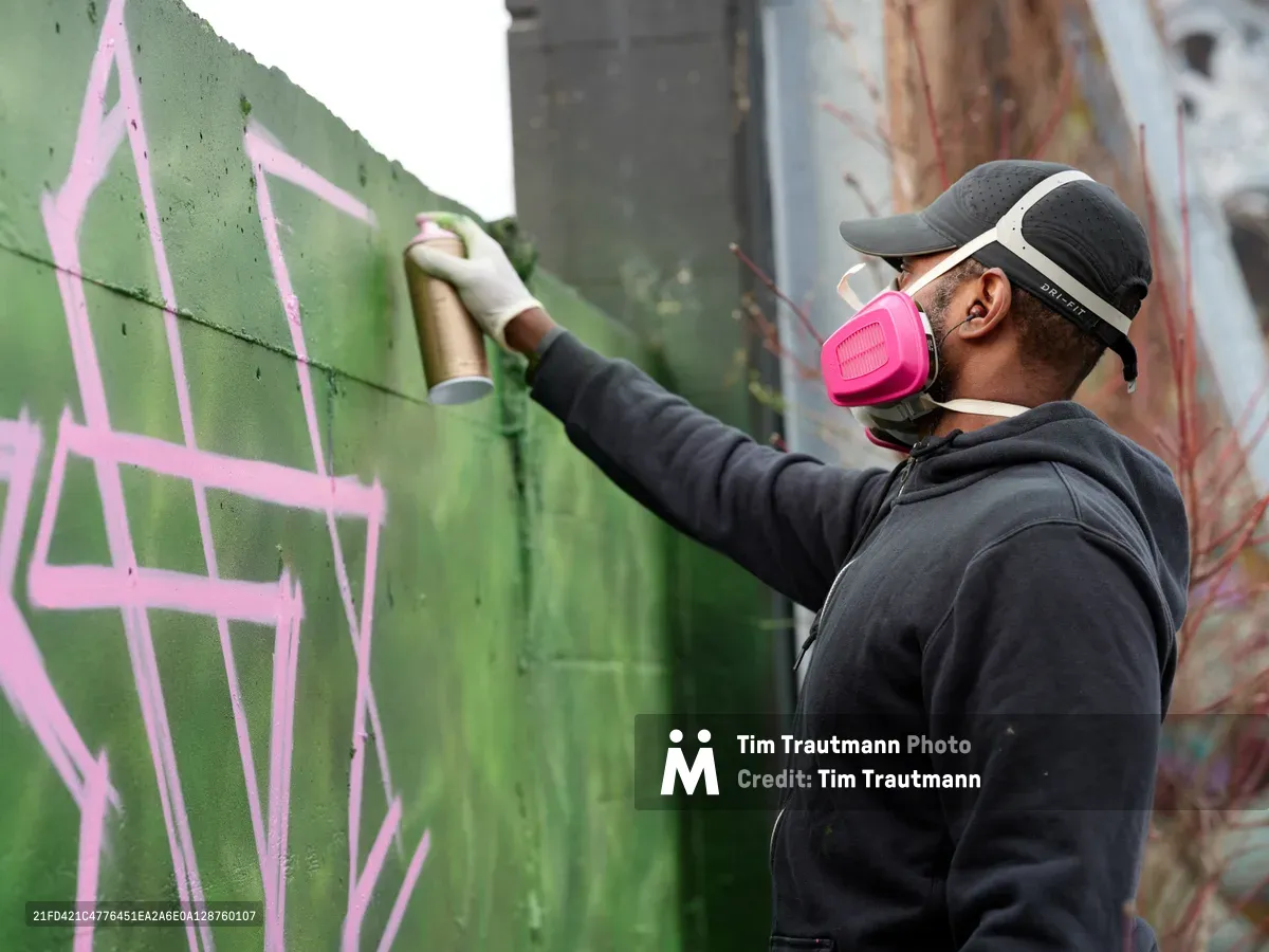 A dedicated street artist wearing a bright pink respirator mask carefully applies green spray paint to a concrete wall, adding bold strokes to an emerging mural. The scene unfolds in the parking lot of Clay Creative in Southeast Portland's Hosford-Abernethy neighborhood, where layers of existing graffiti create a colorful backdrop. Soft daylight illuminates the artist's focused concentration as he extends his arm to reach higher portions of the wall, his black hoodie and cap contrasting against the vibrant pink and green paint.