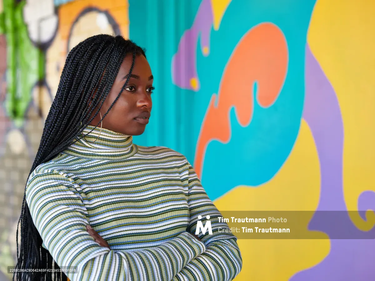 Naomi Likayi stands in quiet contemplation before her newly completed mural on Northeast 15th Avenue in Portland's Alberta district. Her long black braids cascade over a striped turtleneck as she gazes thoughtfully at the explosion of coral, turquoise, and golden hues that dance across the wall behind her. The organic, flowing forms of her street art create a striking contrast against her measured, reflective pose, capturing a moment of artistic satisfaction in this gentrifying corner of Northeast Portland.